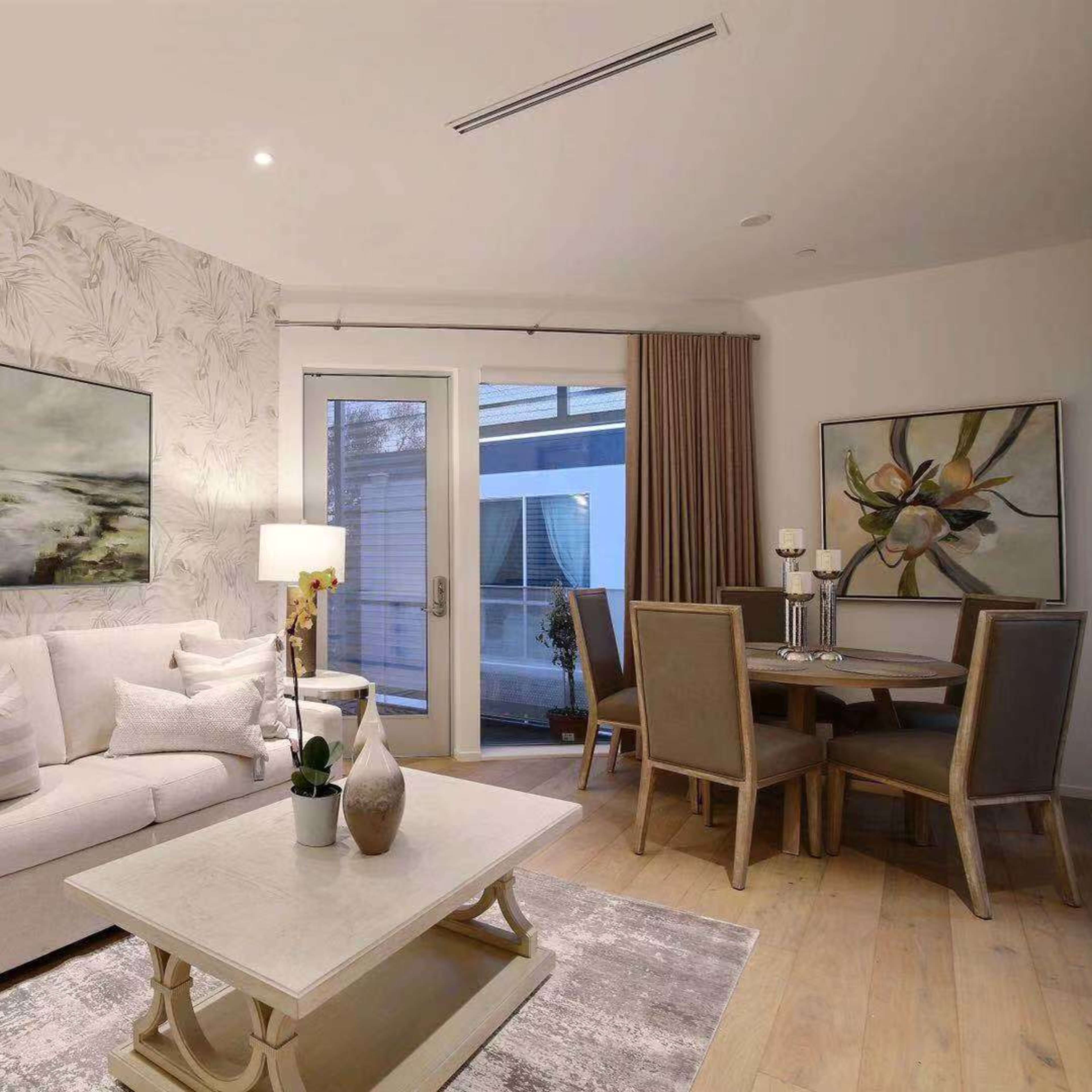 A modern living room with a sofa, decorative lamps, a dining area, and large windows providing natural light.