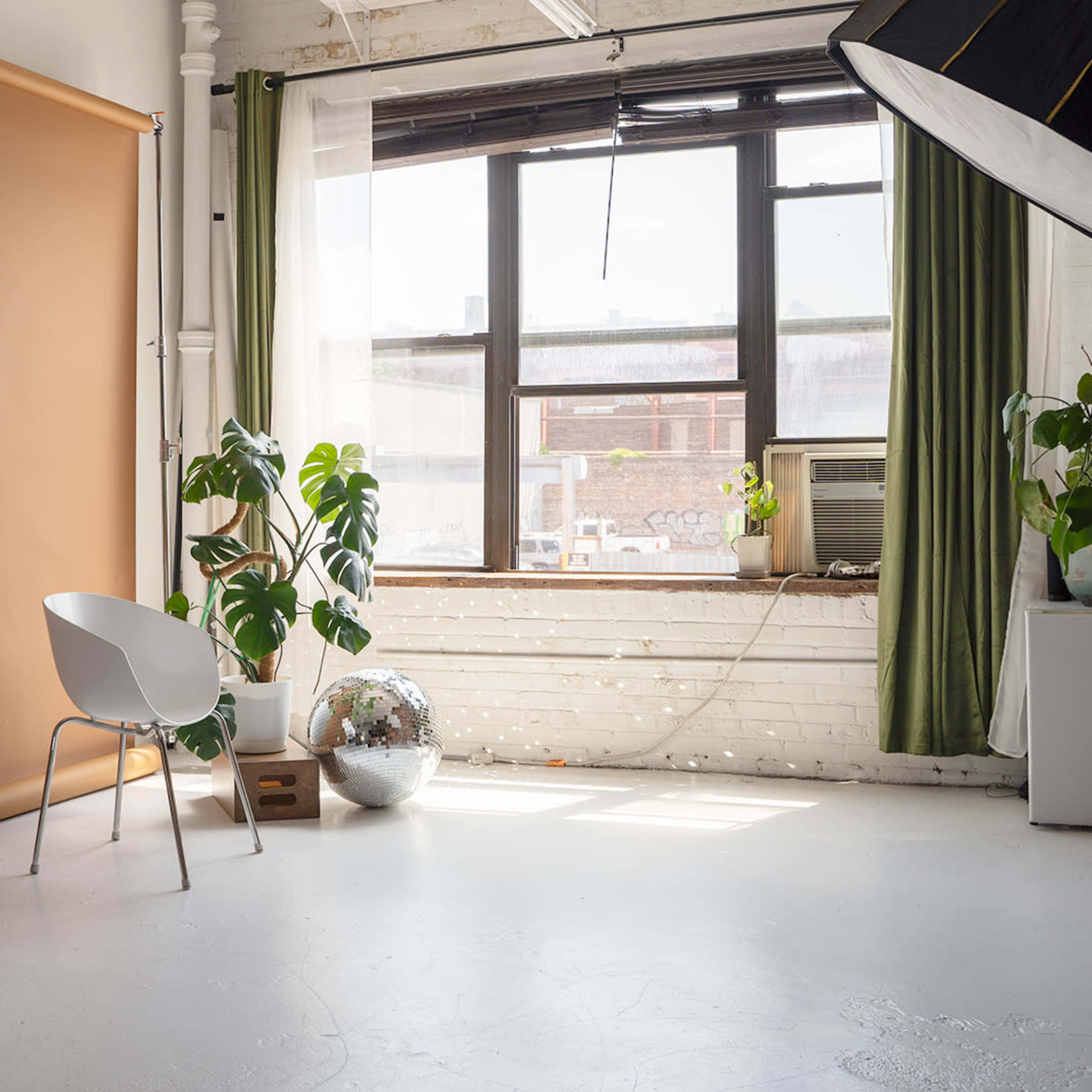 A photography studio featuring a beige backdrop, a white chair, a large window with green curtains, and several indoor plants.