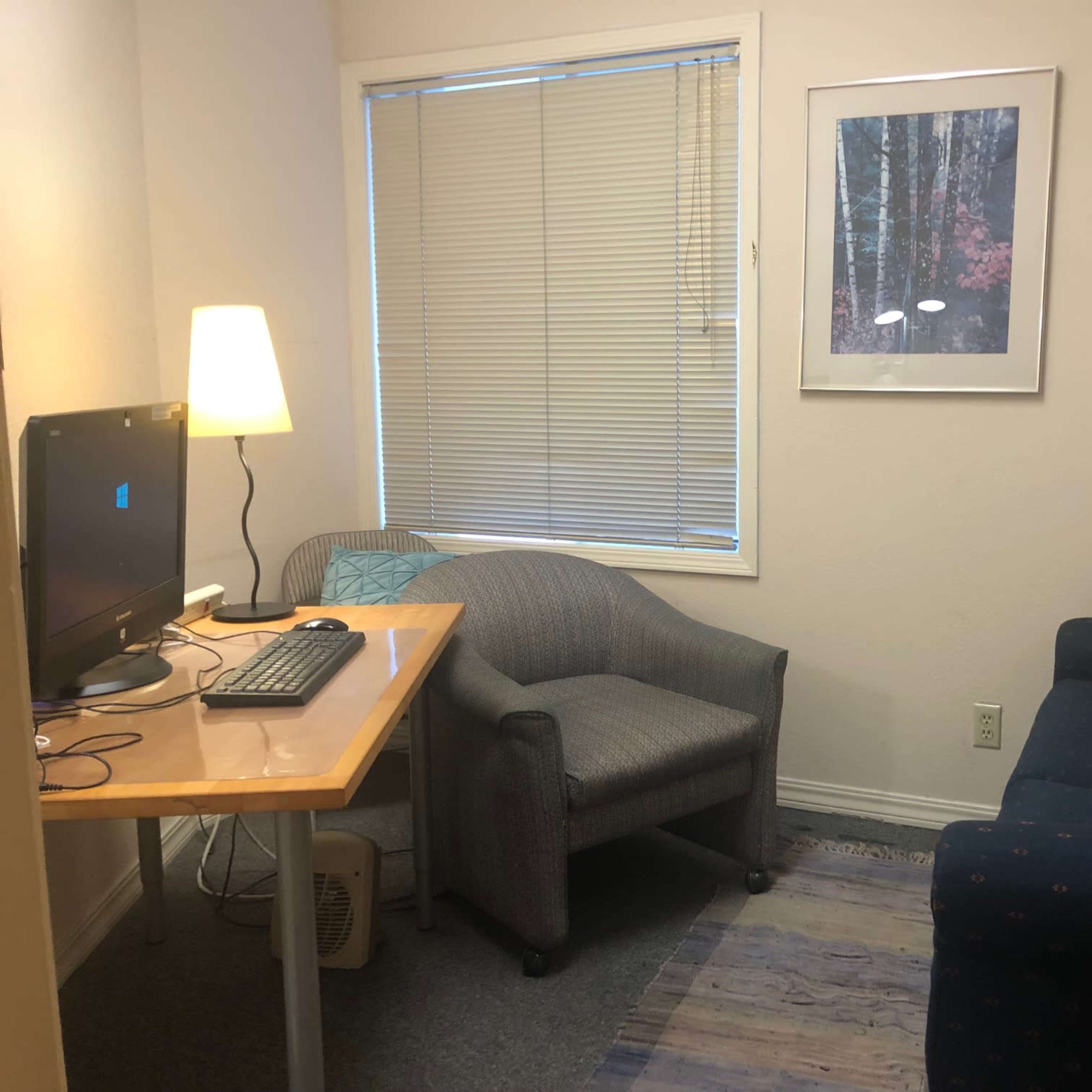 A small room featuring a desk with a computer and a chair, a couch, and a window with blinds.