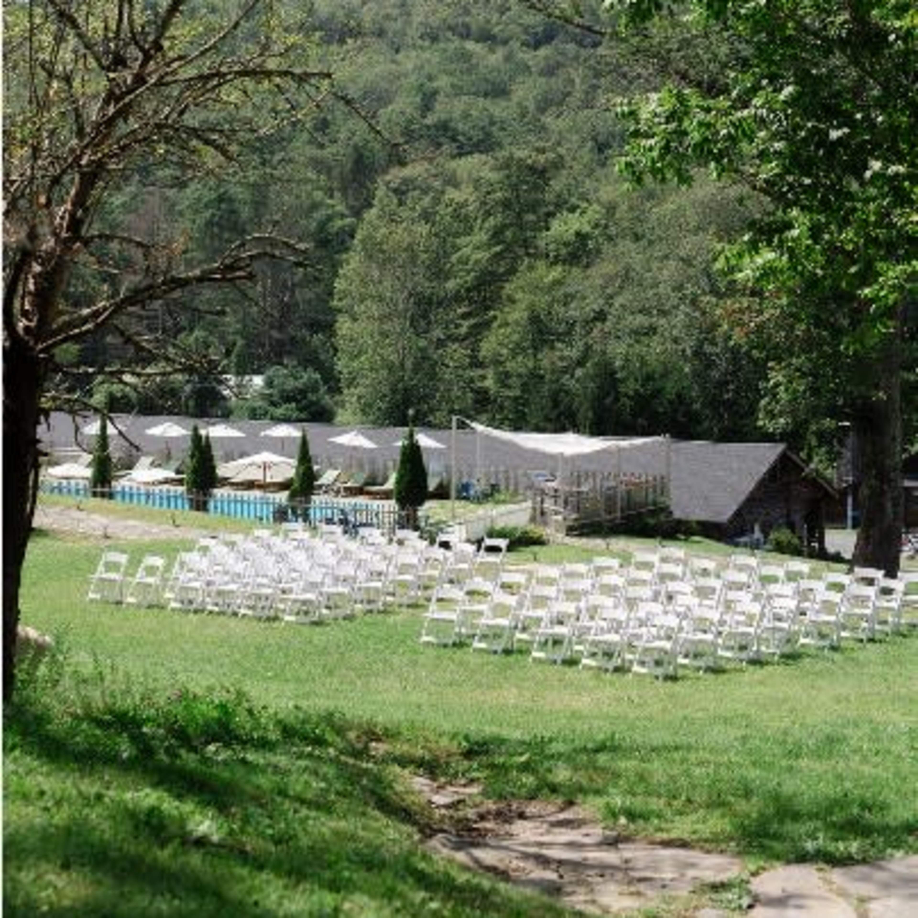 A row of white chairs is set up on a grassy area near a pool and a forested hillside, preparing for an outdoor event.