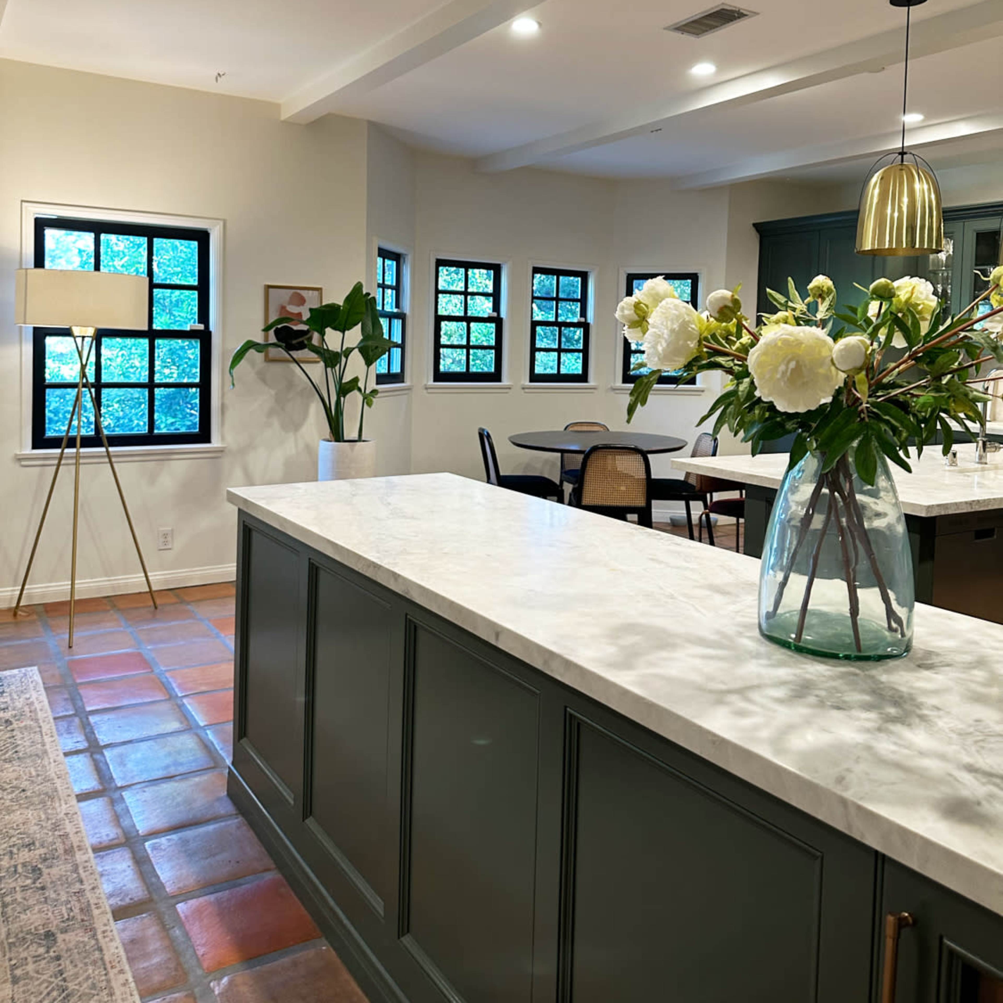 A modern kitchen features a marble countertop, green cabinetry, and large windows with potted plants and a dining area in the background.
