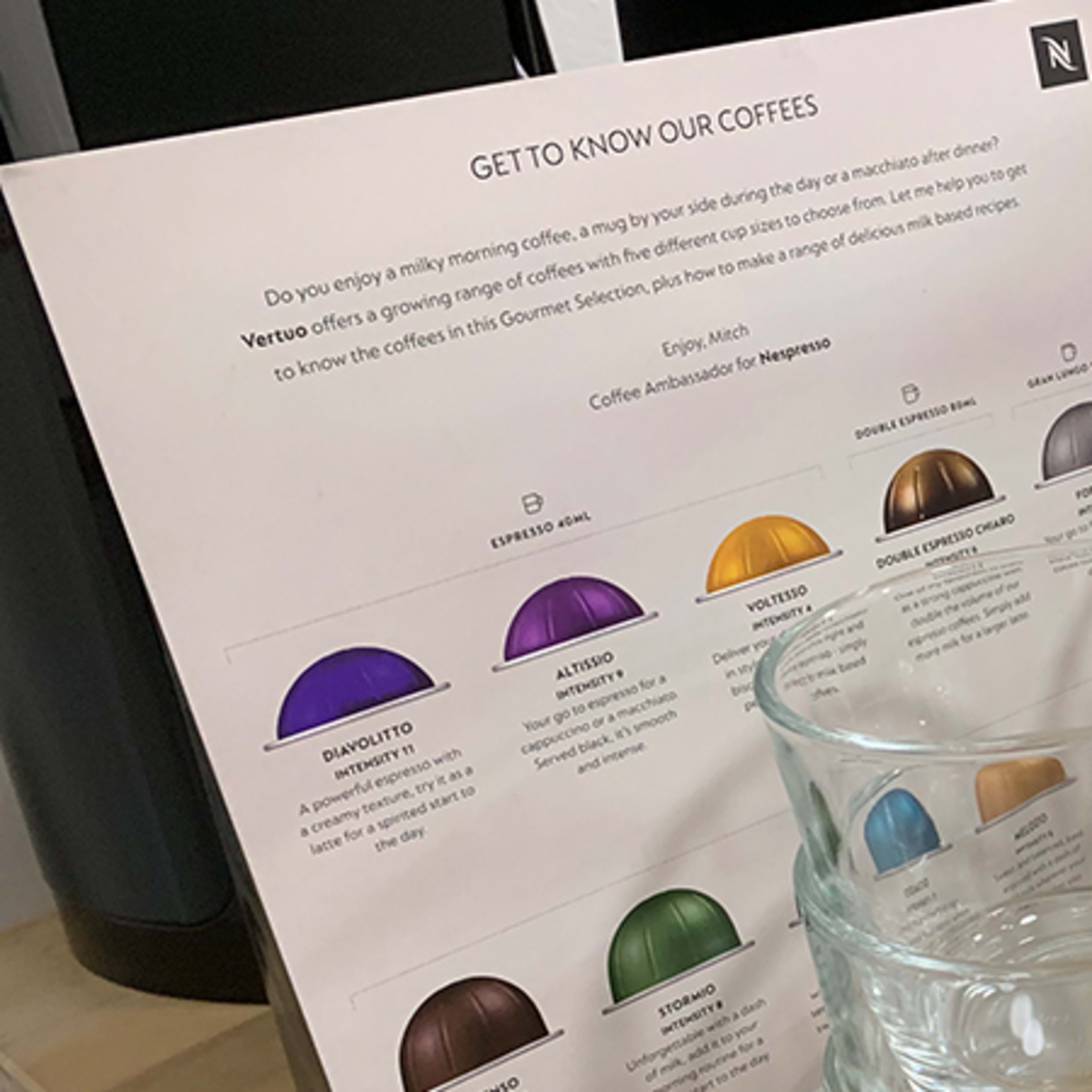 A coffee station features a brochure about different coffee blends alongside several clear glass mugs and a coffee machine.