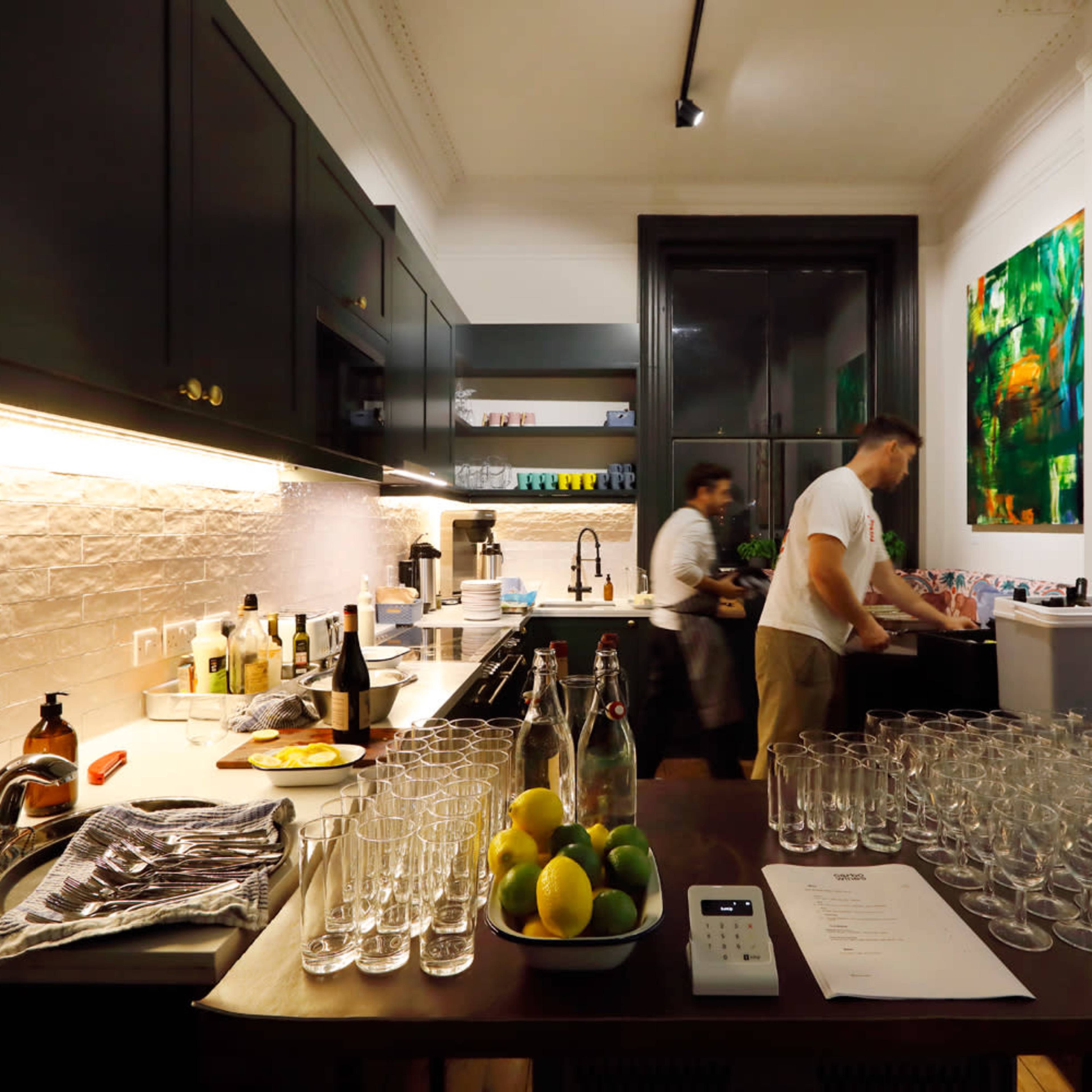Two people are preparing drinks in a modern kitchen with a long row of clear glasses and various ingredients on the counter.