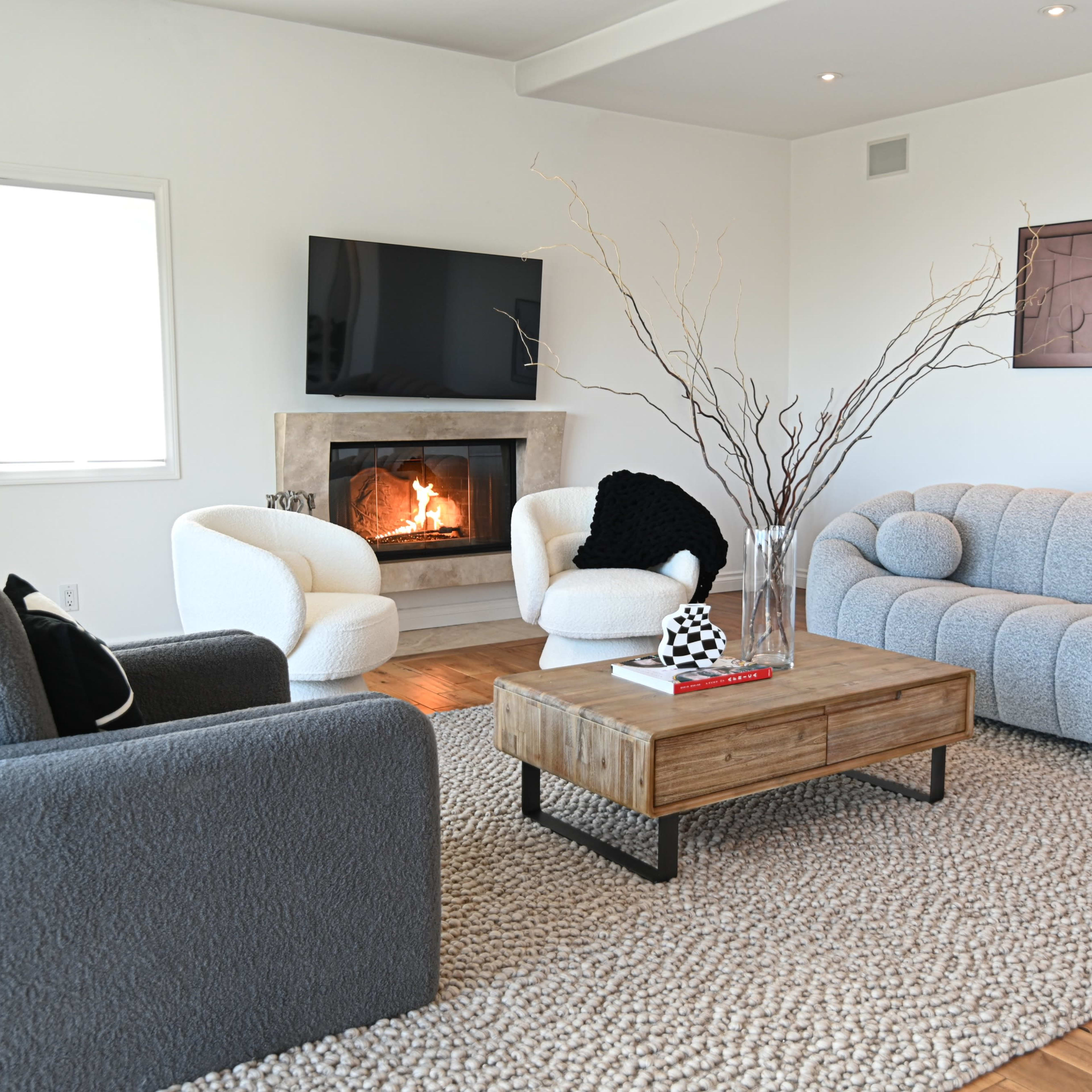 The living room features a modern design with a plush gray sofa, two armchairs, a wooden coffee table, and a fireplace set against a light-colored wall.