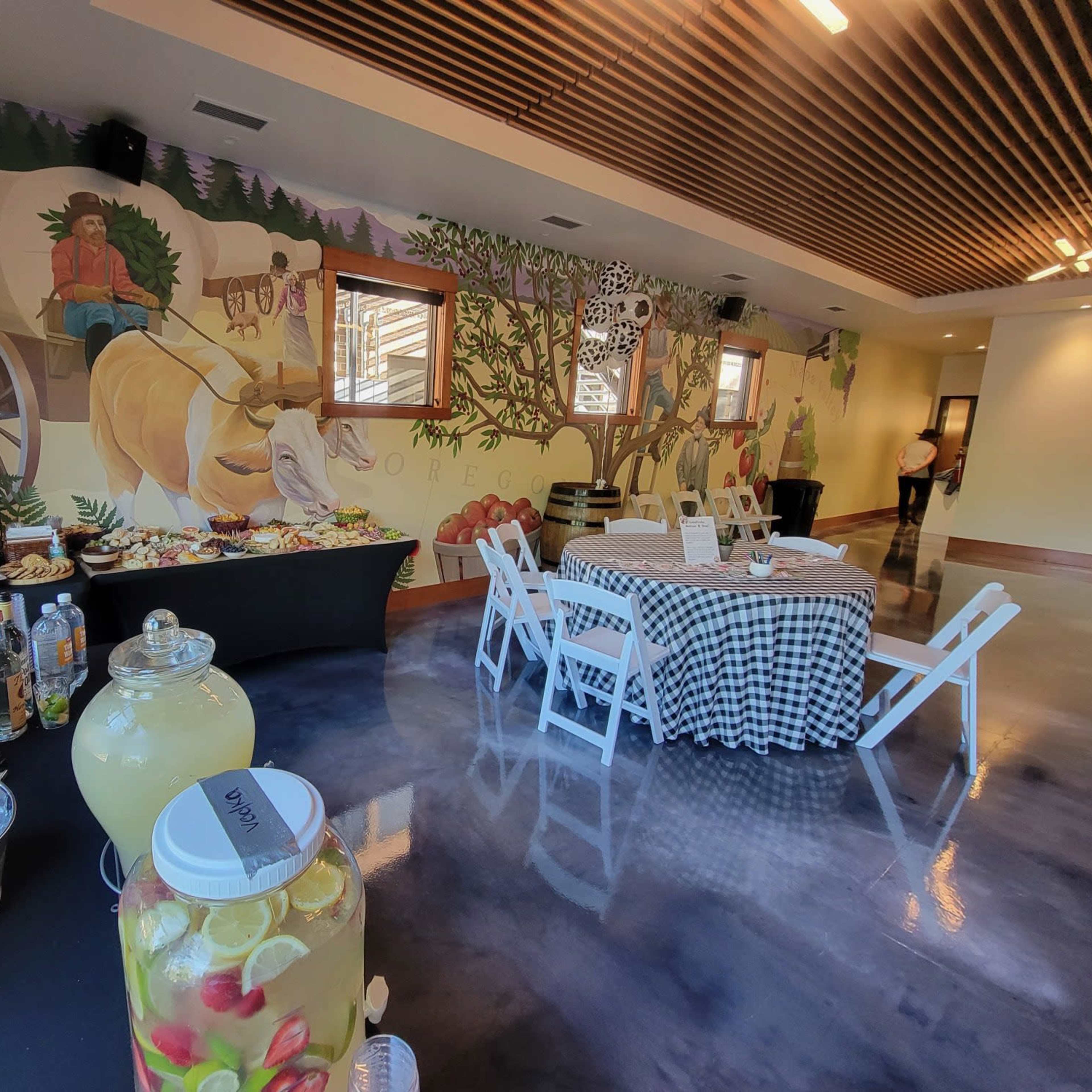 A spacious indoor event space with a mural depicting a farmer, a table laden with food, and a drink station featuring a large glass jar of lemonade.