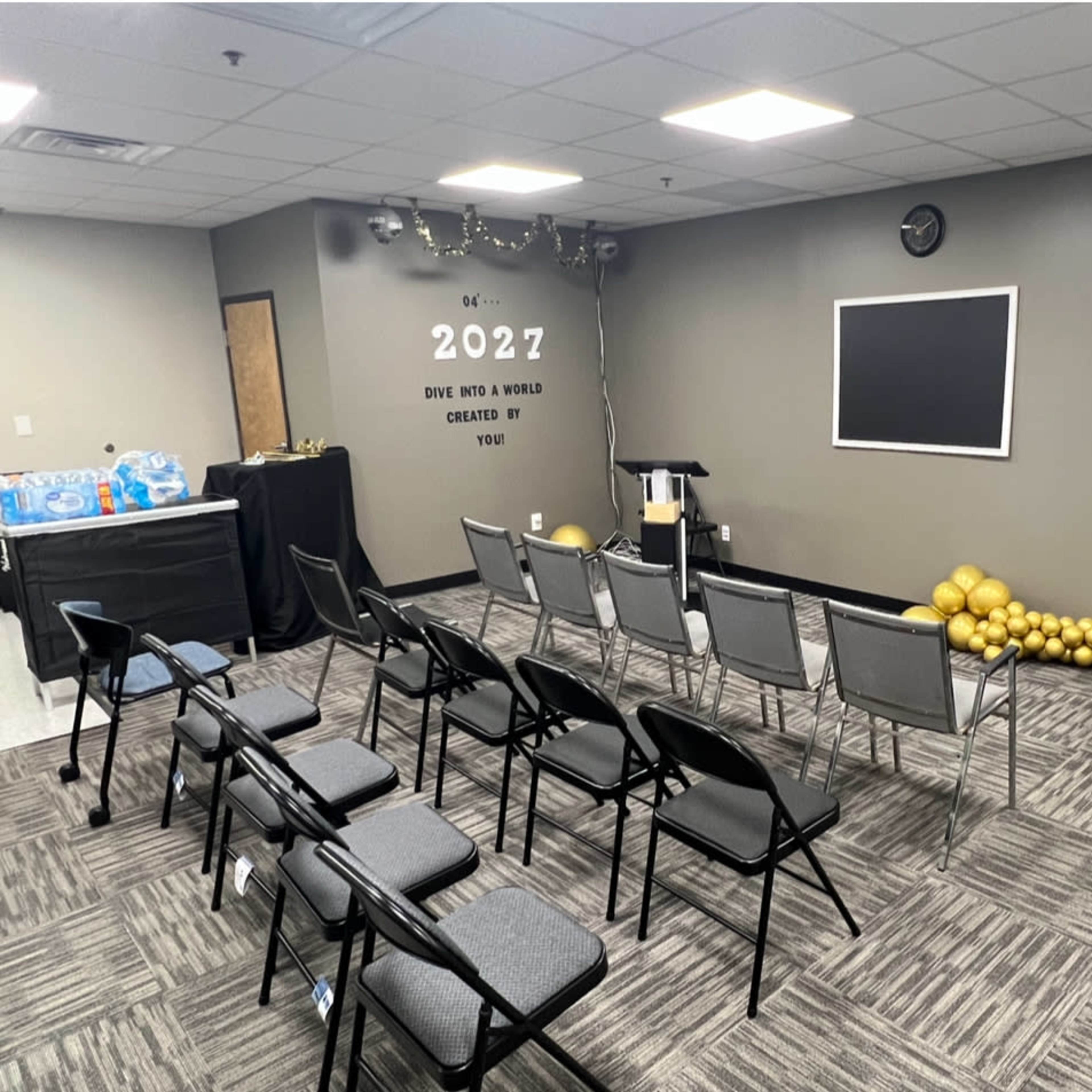A conference room set up with several gray chairs arranged in rows, a black podium at the front, and a wall featuring decorative elements including a chalkboard and posters.
