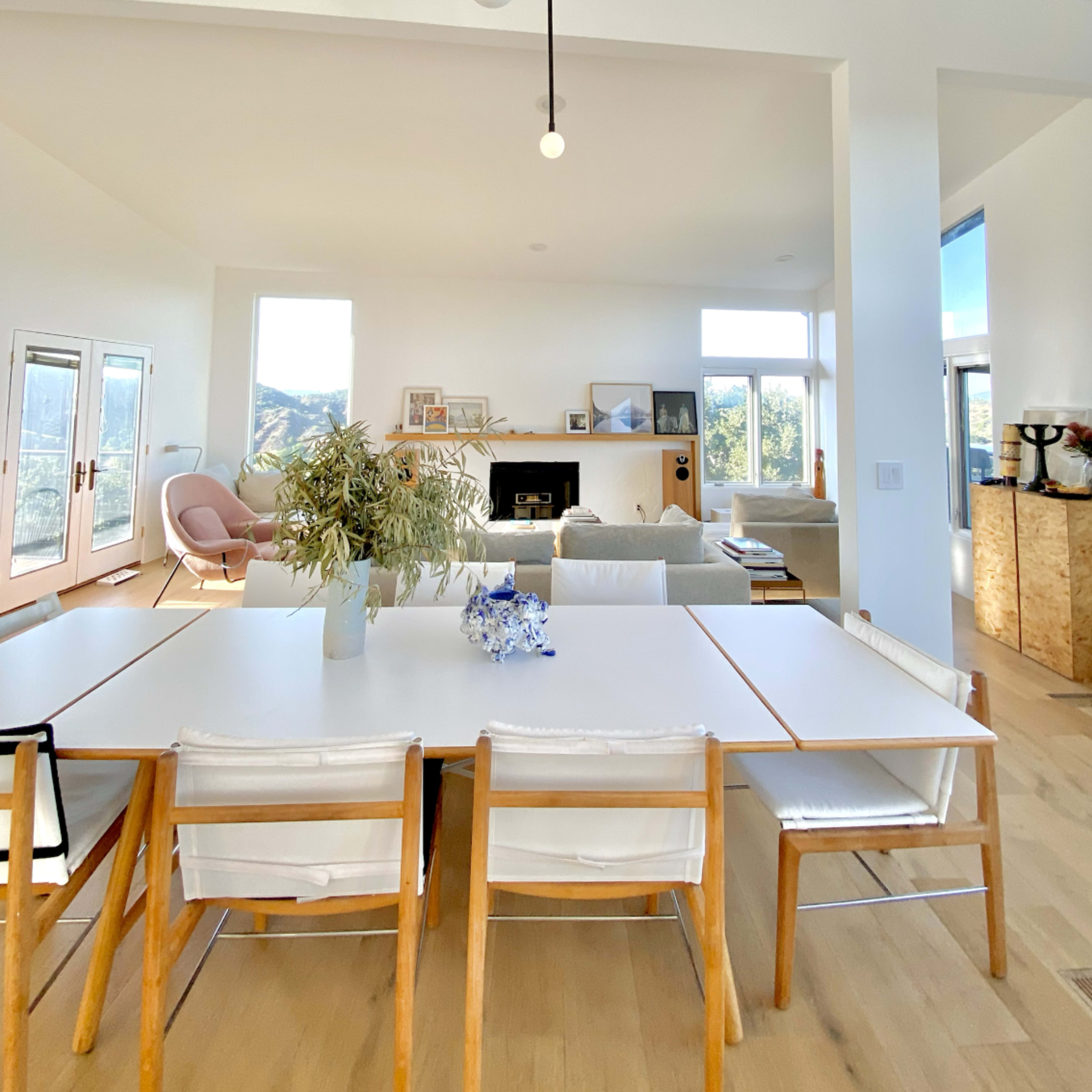 A bright, modern dining area with a large white table and wooden chairs, leading into a spacious living room with a fireplace and large windows.