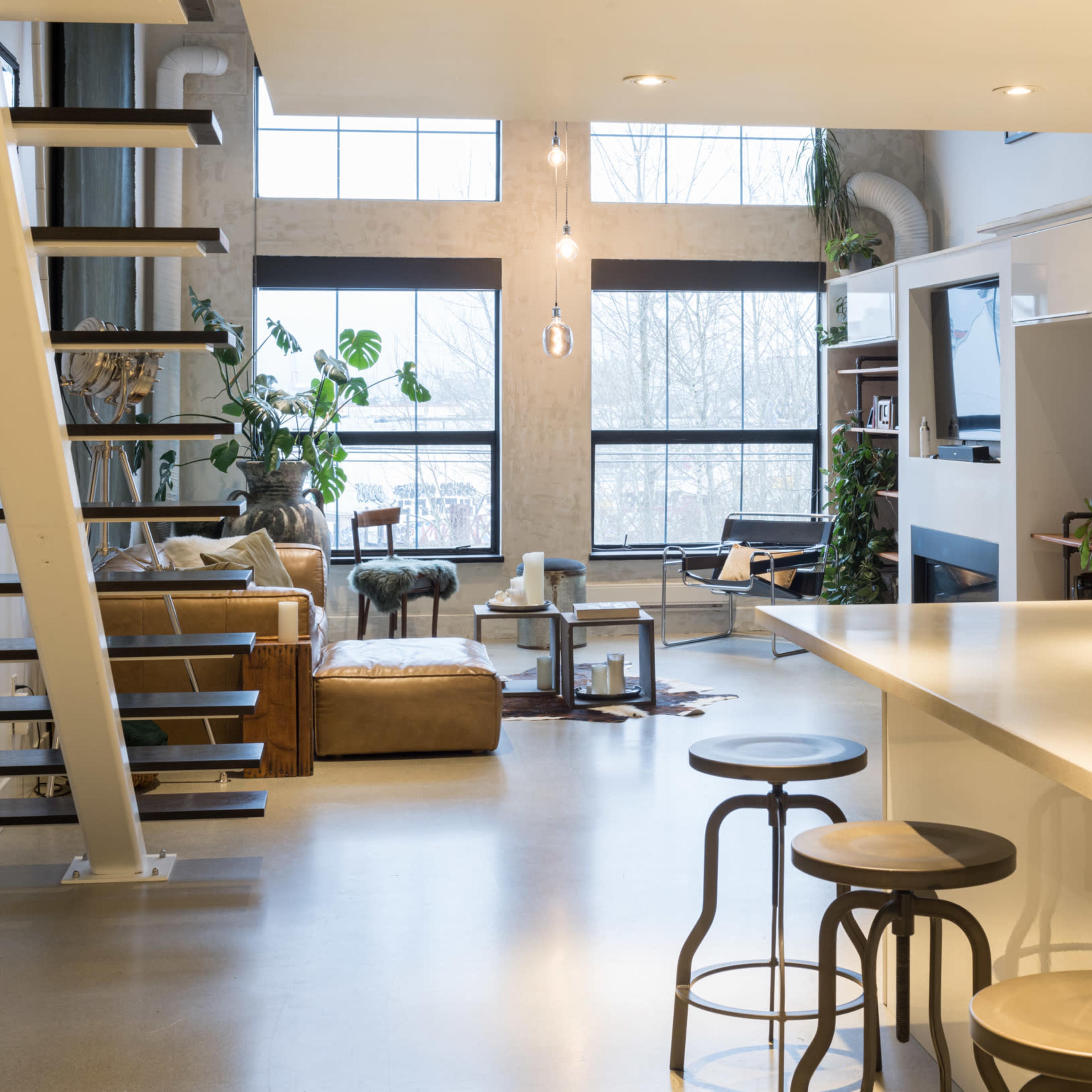 A modern open-concept living space features a kitchen with a central island, bar stools, and a staircase leading to an upper level, while a cozy seating area is visible through large windows filled with plants.