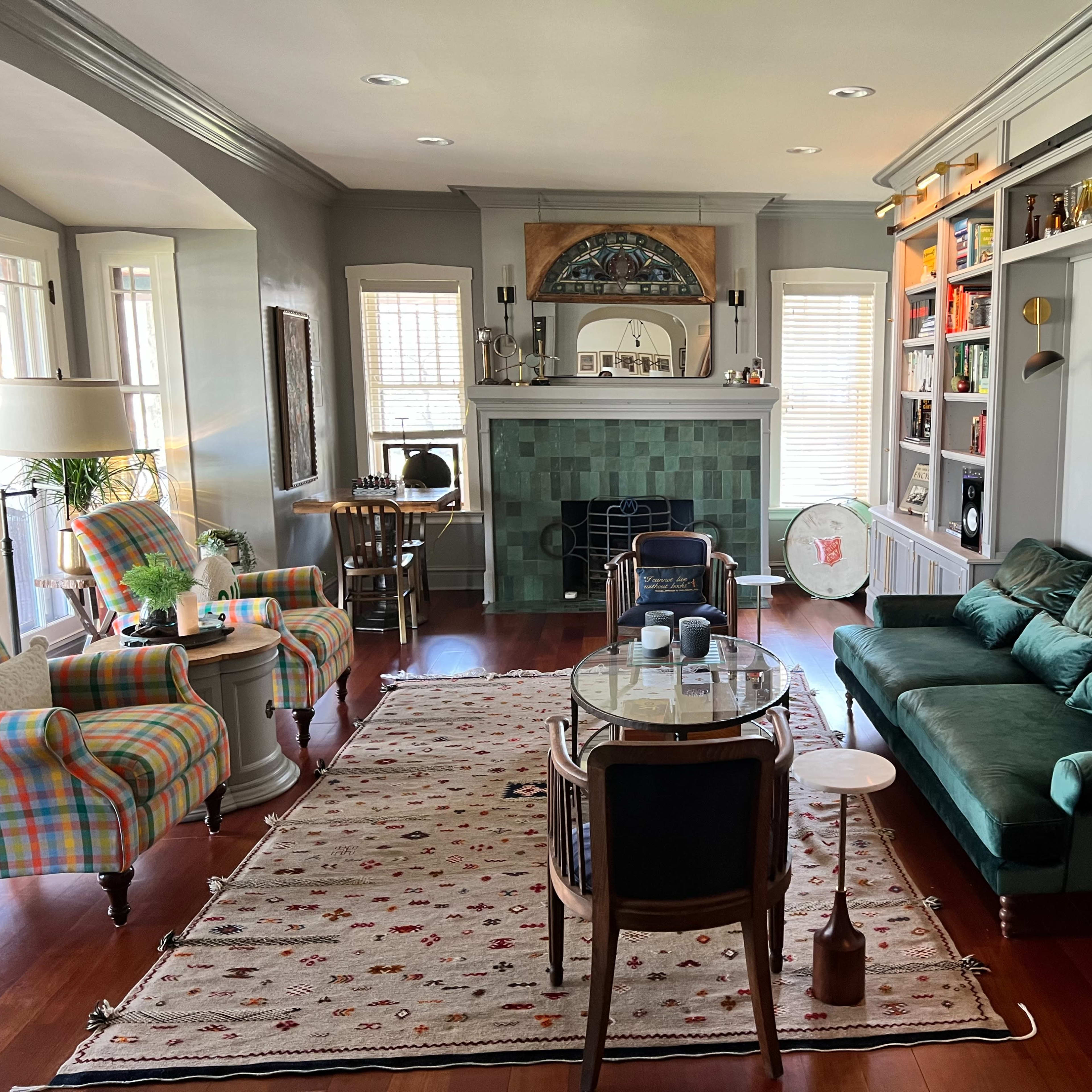 The living room features a green sofa, a patterned armchair, and a decorative fireplace, with shelves displaying various items and a large area rug on the wooden floor.