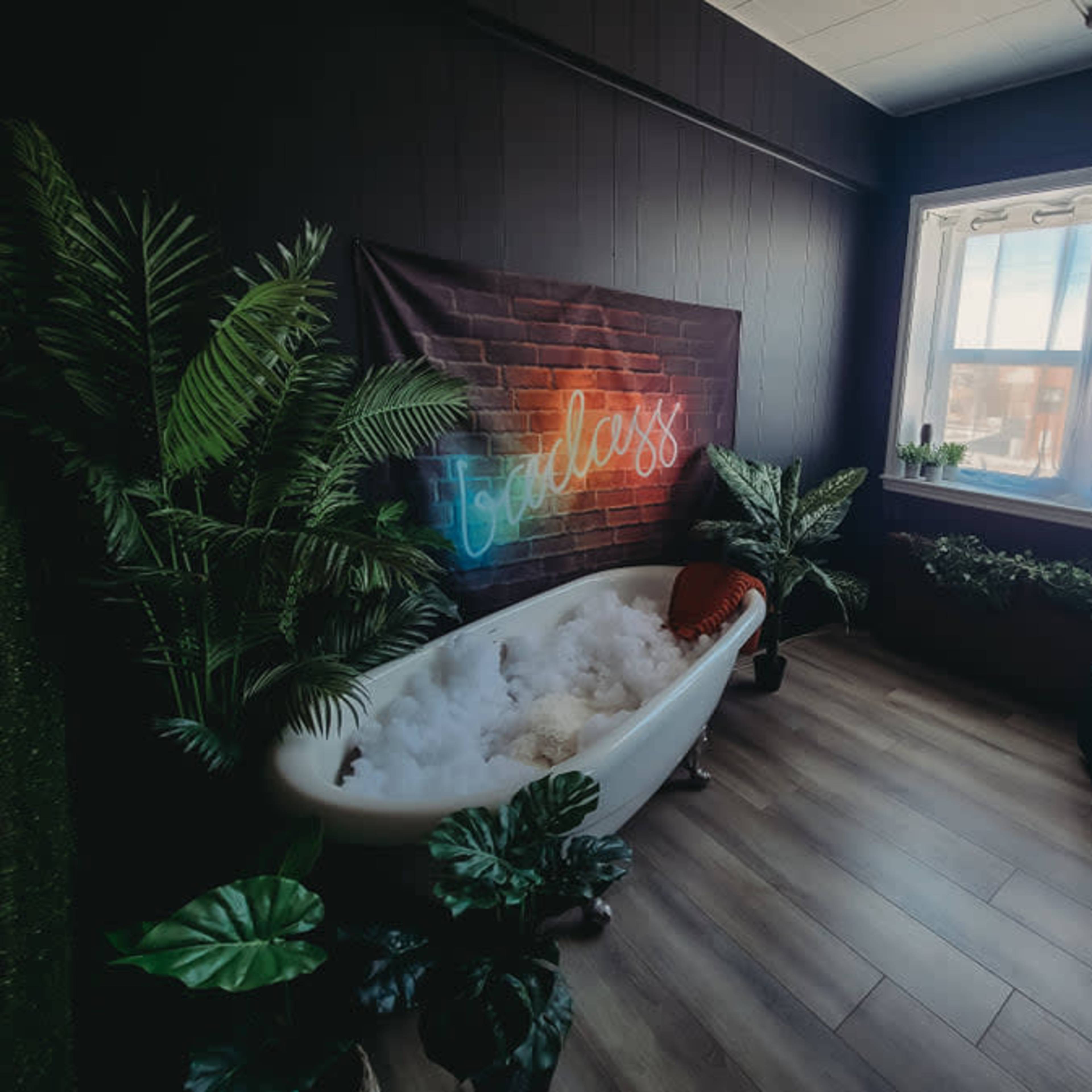 A small, stylish bathroom with a vintage bathtub filled with bubbles, surrounded by indoor plants and a large window letting in natural light.