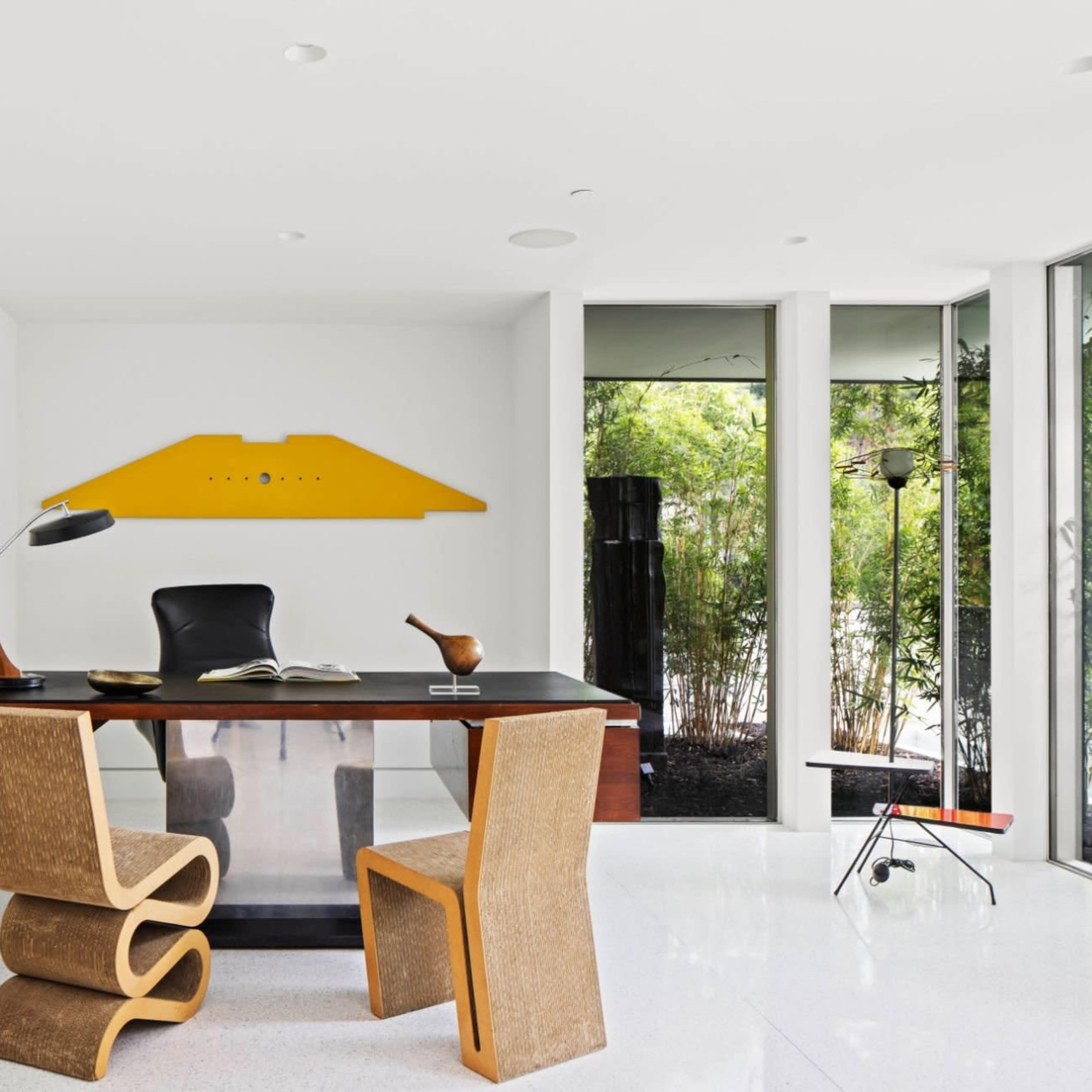 A modern office space featuring a desk, two unique chairs, a bookshelf, and large windows overlooking greenery.