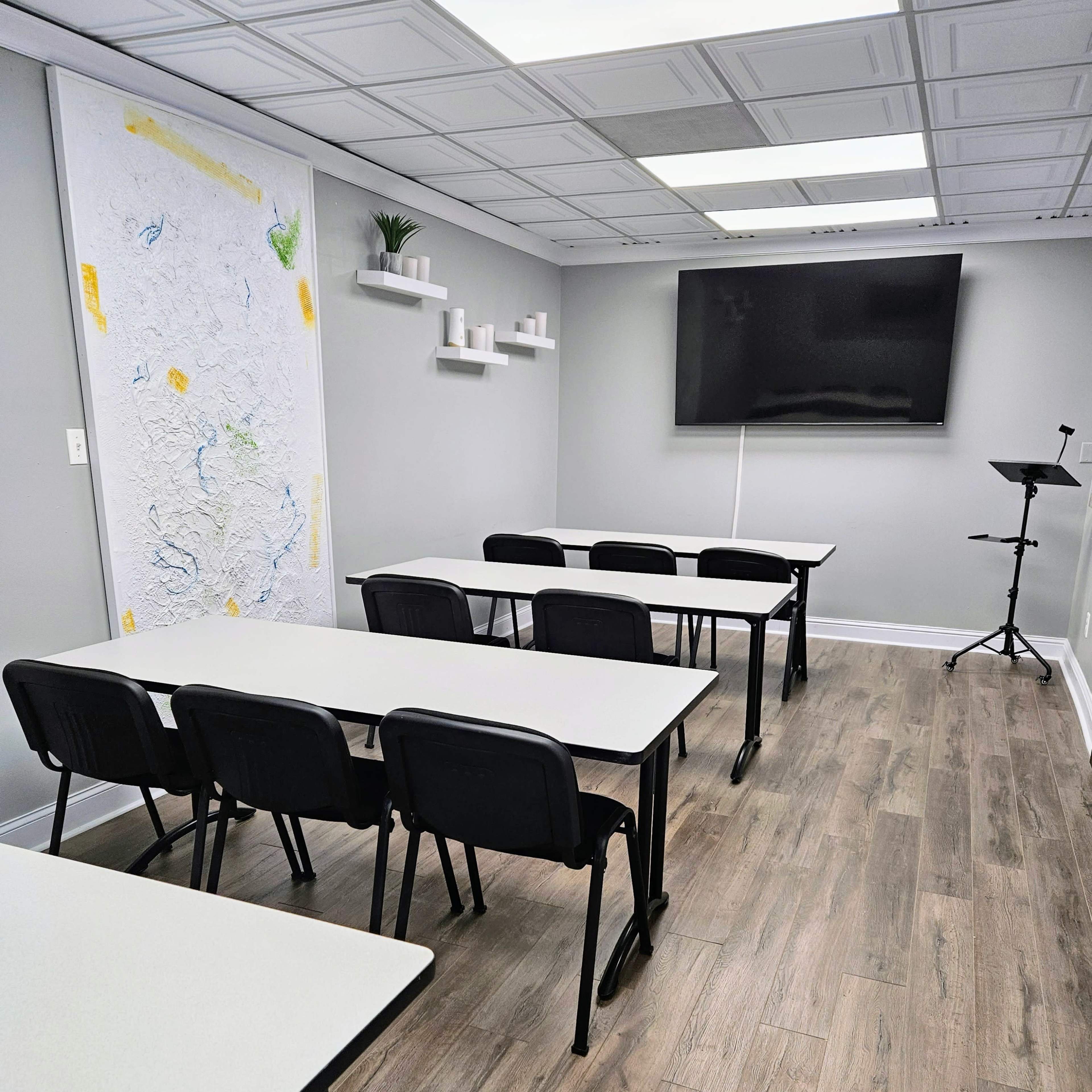 A modern classroom or meeting room with several tables and chairs arranged in rows, a large screen on the wall, and a whiteboard with a map sketch.