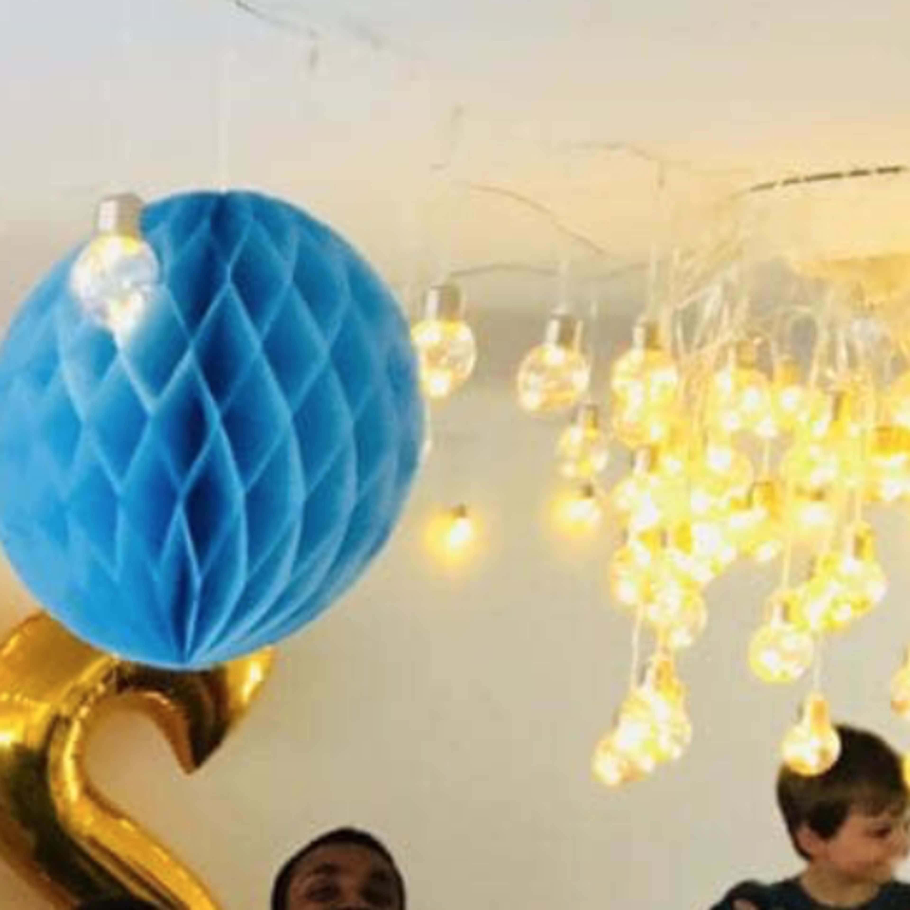A decorated room with golden balloons spelling "PRIS," colorful paper decorations, and numerous hanging light bulbs.
