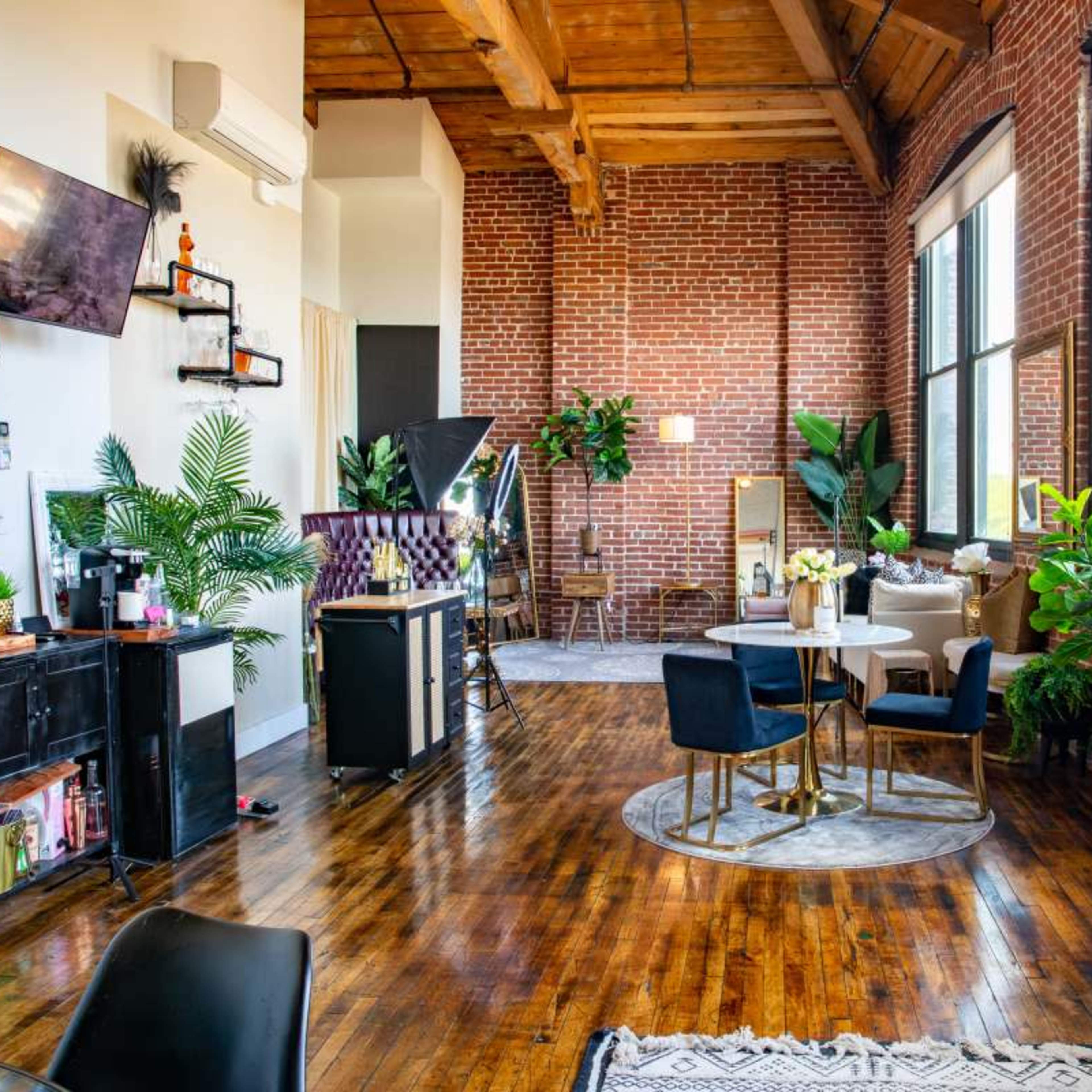 A spacious loft-style room with exposed brick walls, wooden beams, and various plants, featuring a seating area with chairs and a table.