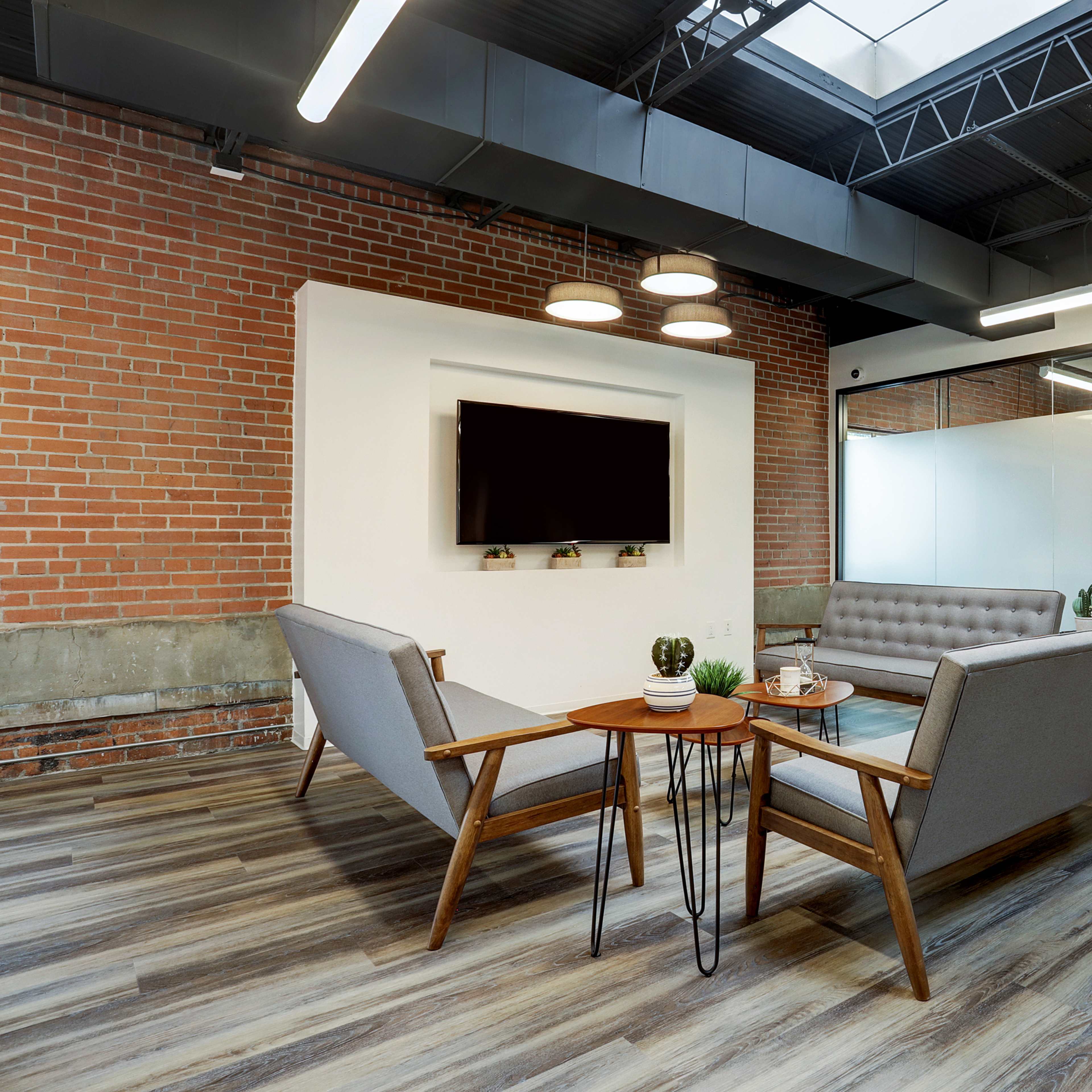 A modern office lounge area with a brick wall, two gray sofas facing a wooden table, and a television mounted on a white wall.