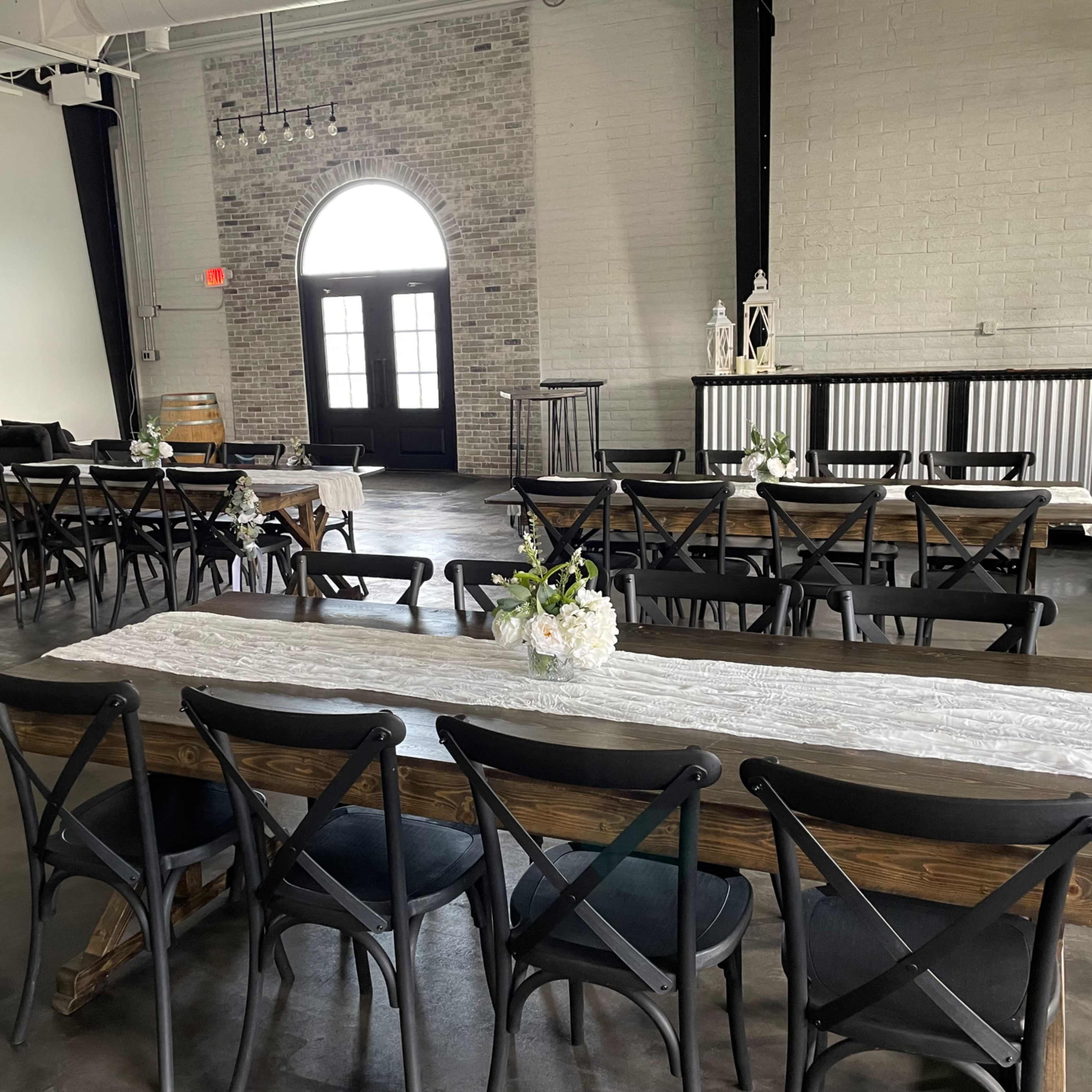 An indoor event space featuring several long wooden tables arranged with chairs and floral centerpieces, with large windows and a bar area in the background.