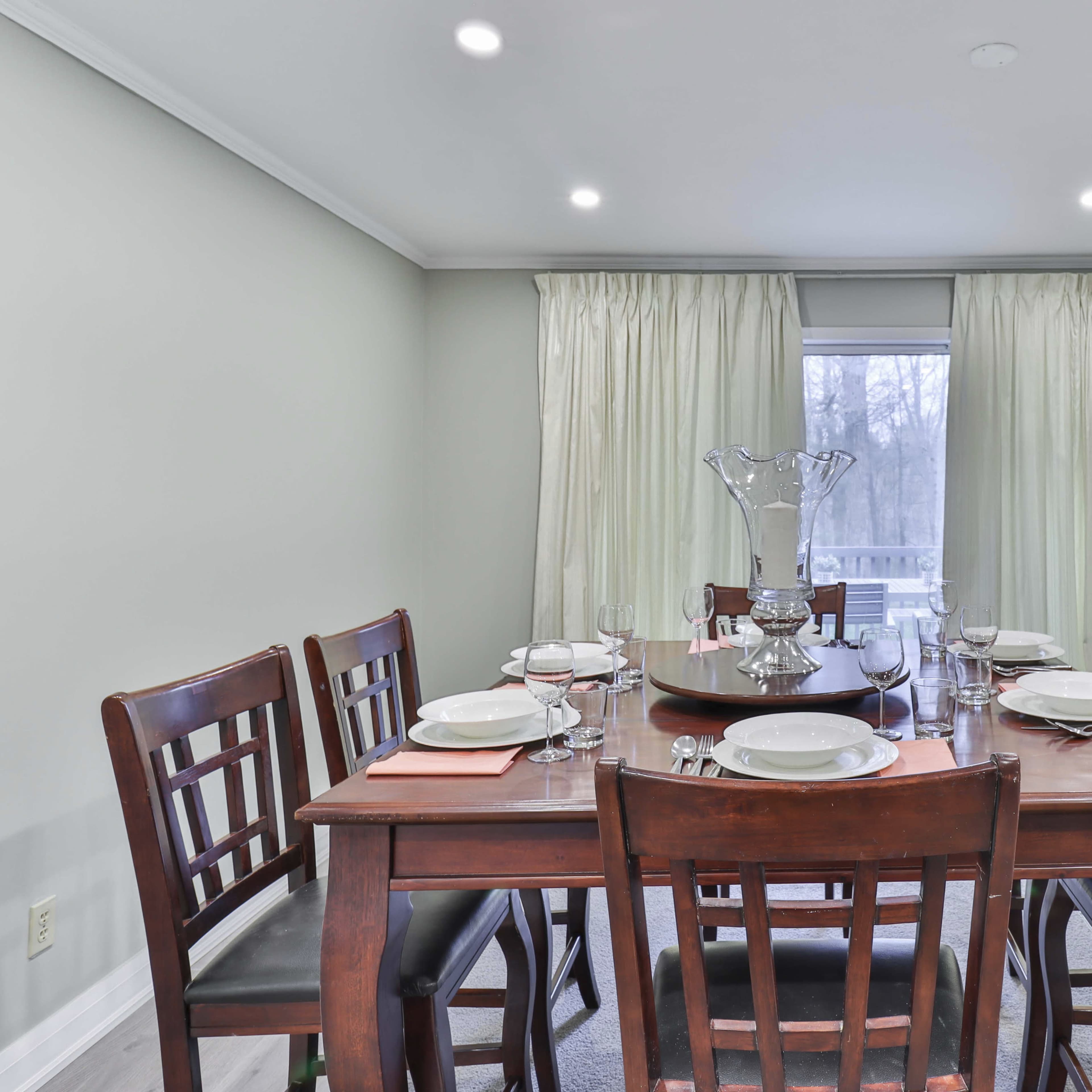 A dining room features a wooden table set for eight with plates, glasses, and a centerpiece, surrounded by chairs and large windows draped with curtains.