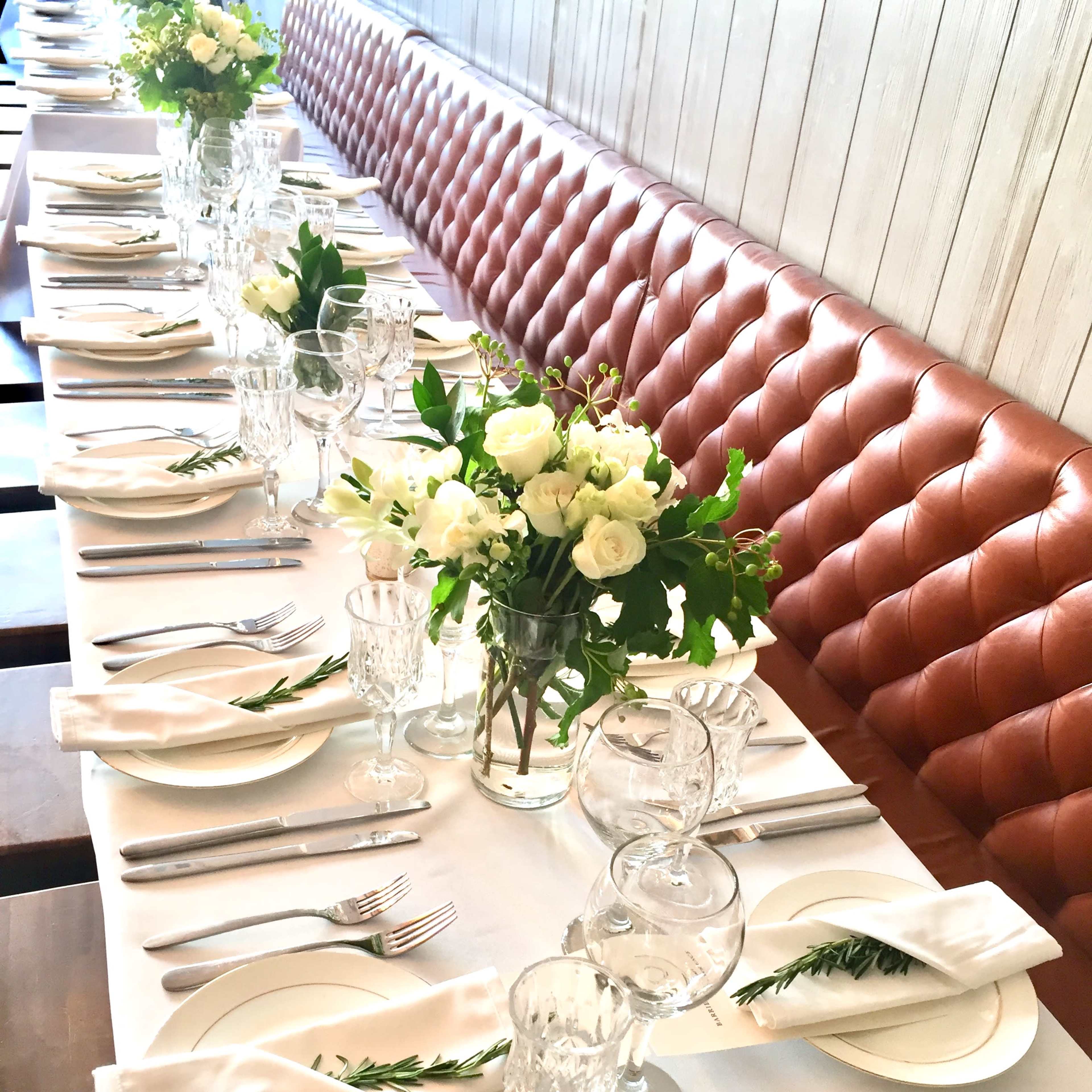 A long dining table is elegantly set with white plates, glassware, and floral centerpieces, arranged along a leather-upholstered bench.