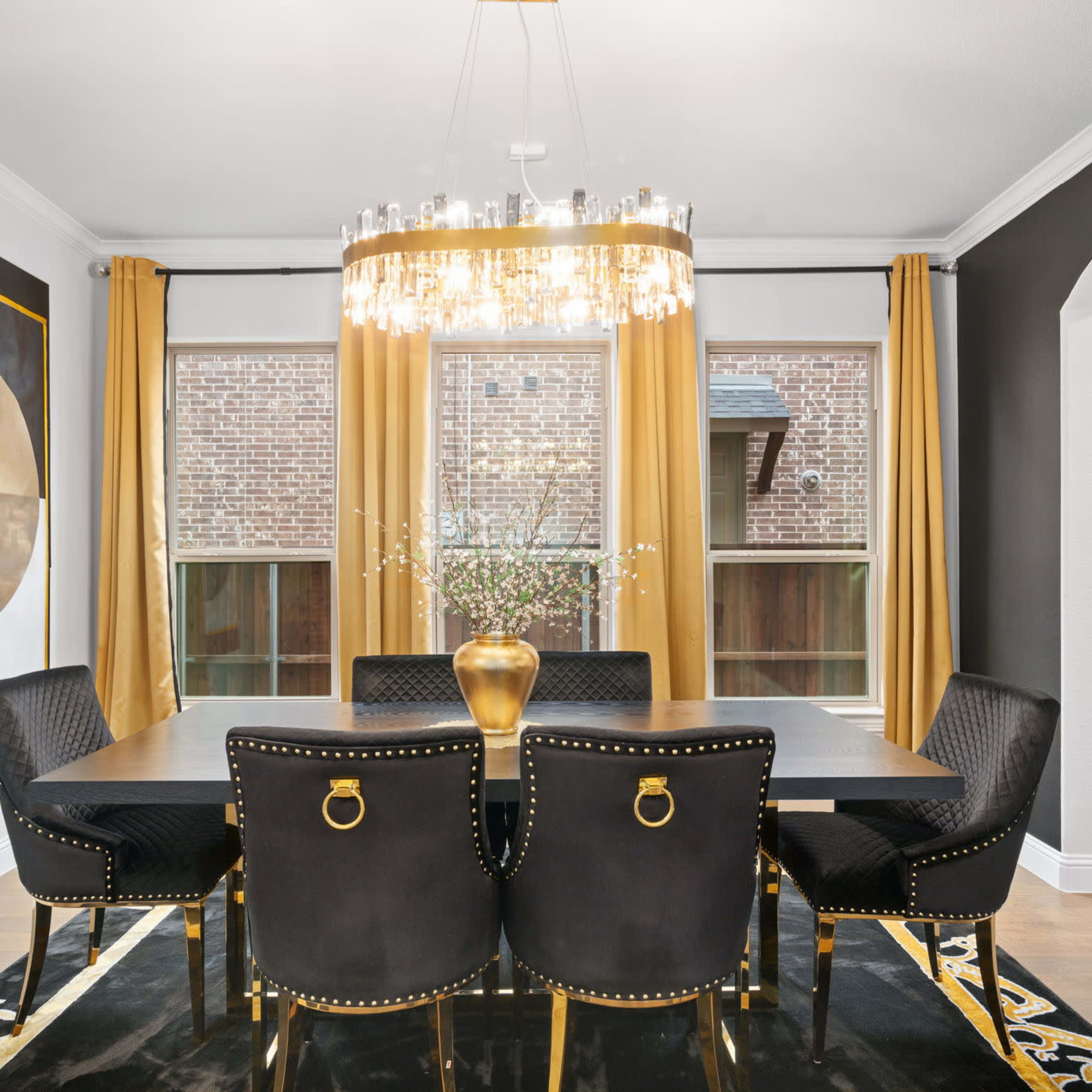 A modern dining room features a large table surrounded by black chairs, a chandelier overhead, and large windows dressed with golden curtains.