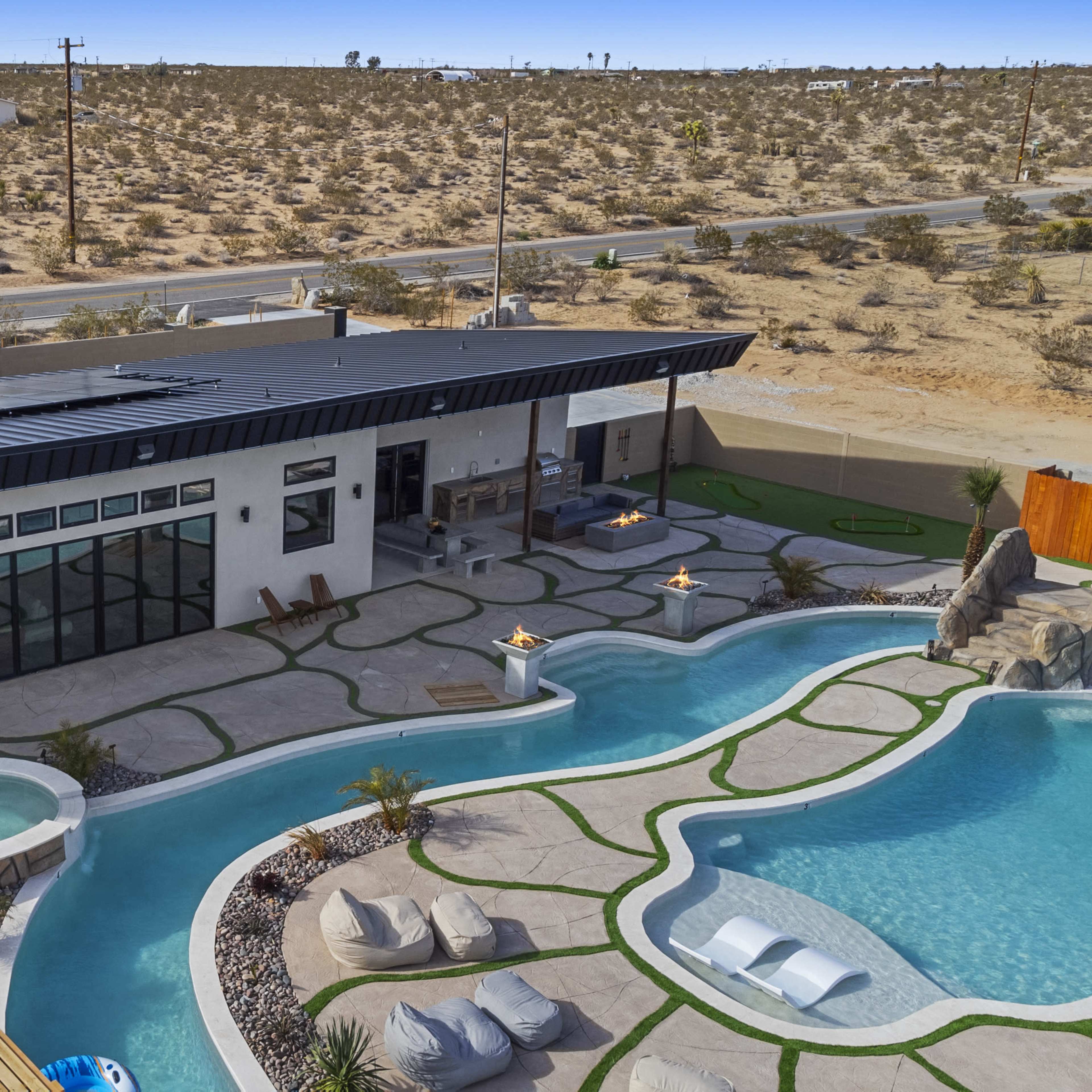 A modern house with large windows and a black roof, surrounded by a landscaped pool area and desert terrain.
