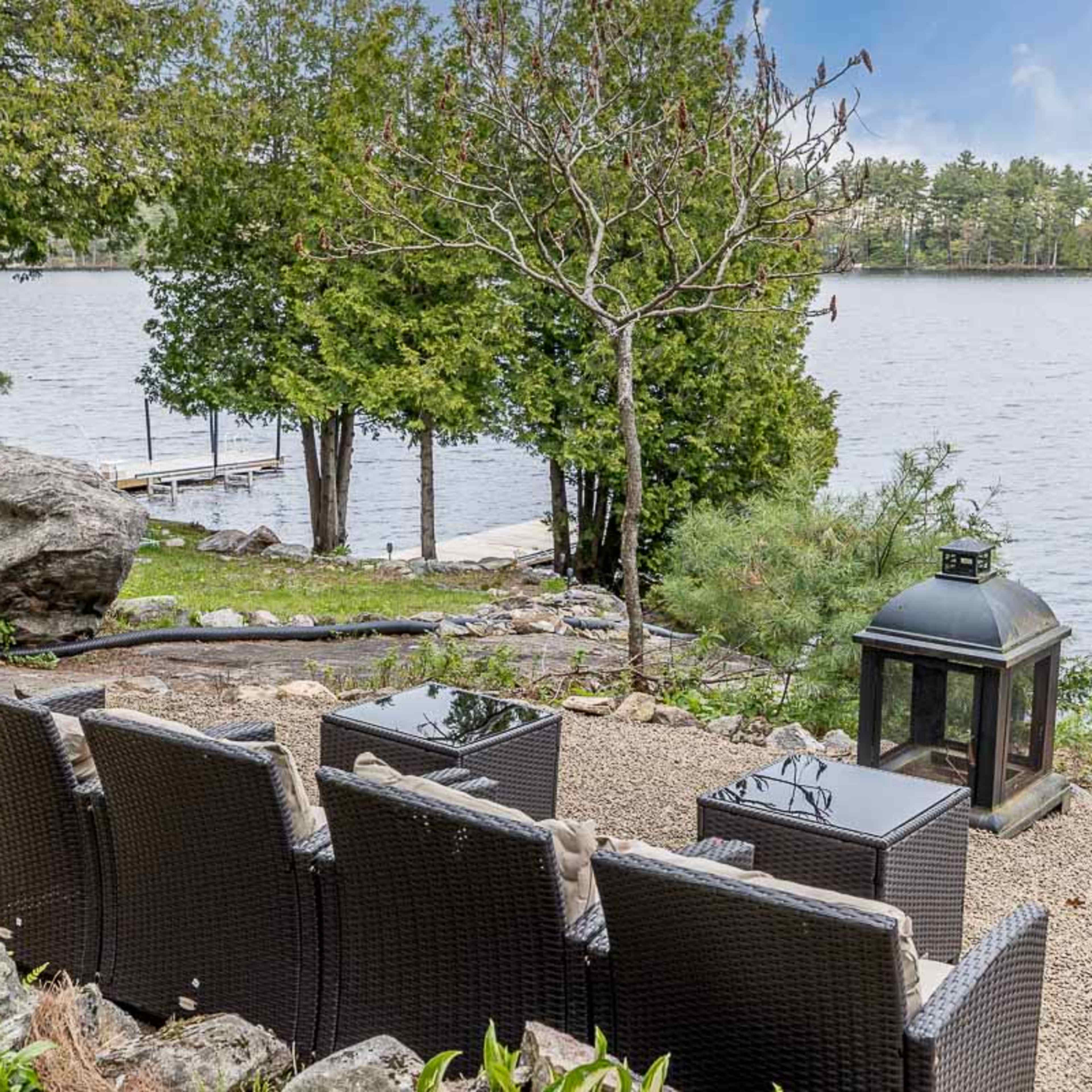 A lakeside seating area with four wicker chairs, small tables, a lantern, and a view of the water and trees.