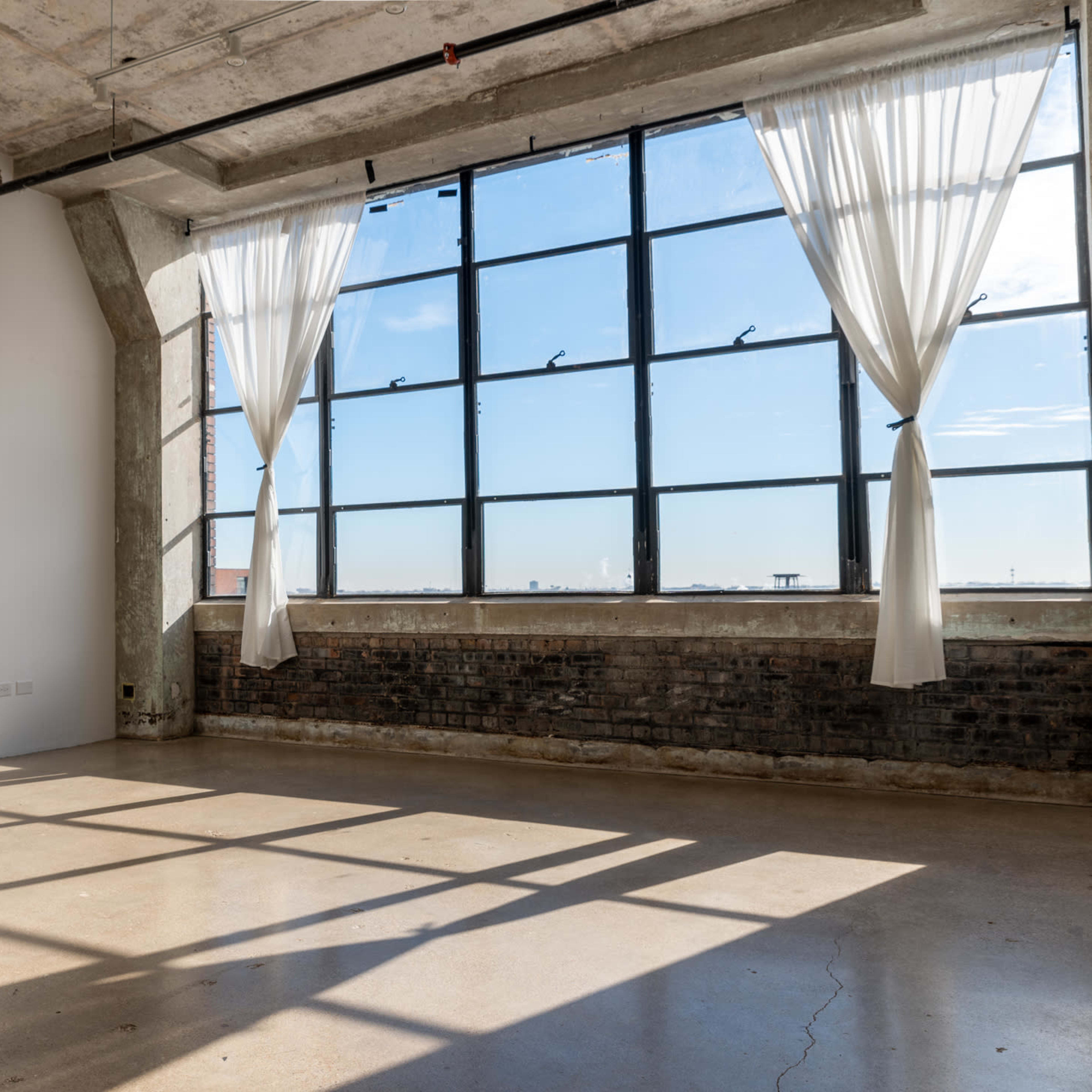 A spacious room with large industrial windows covered by sheer white curtains, casting shadows on a polished concrete floor.