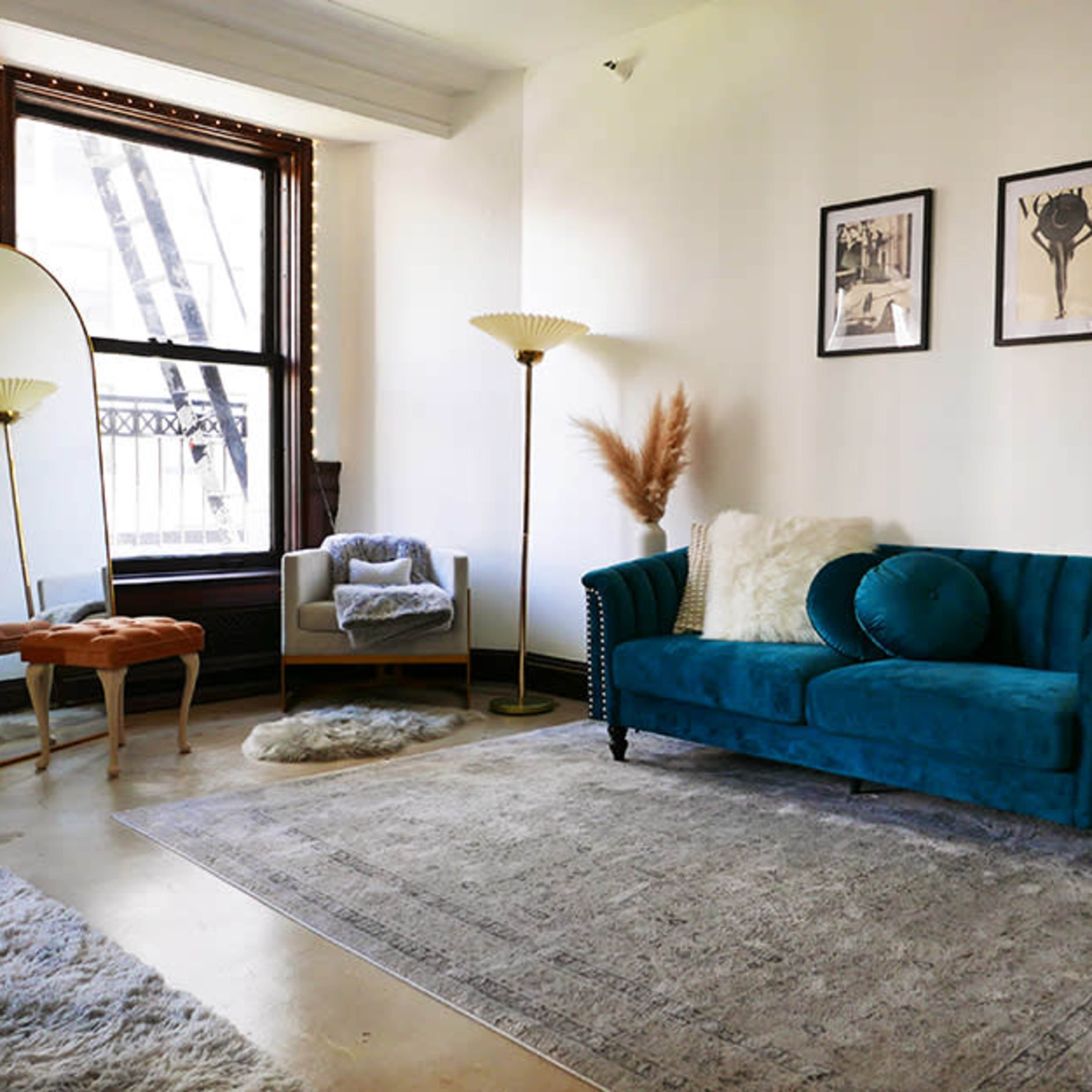 A well-decorated living room featuring a teal sofa, a large mirror, floor lamps, and decorative plants arranged around a plush area rug.
