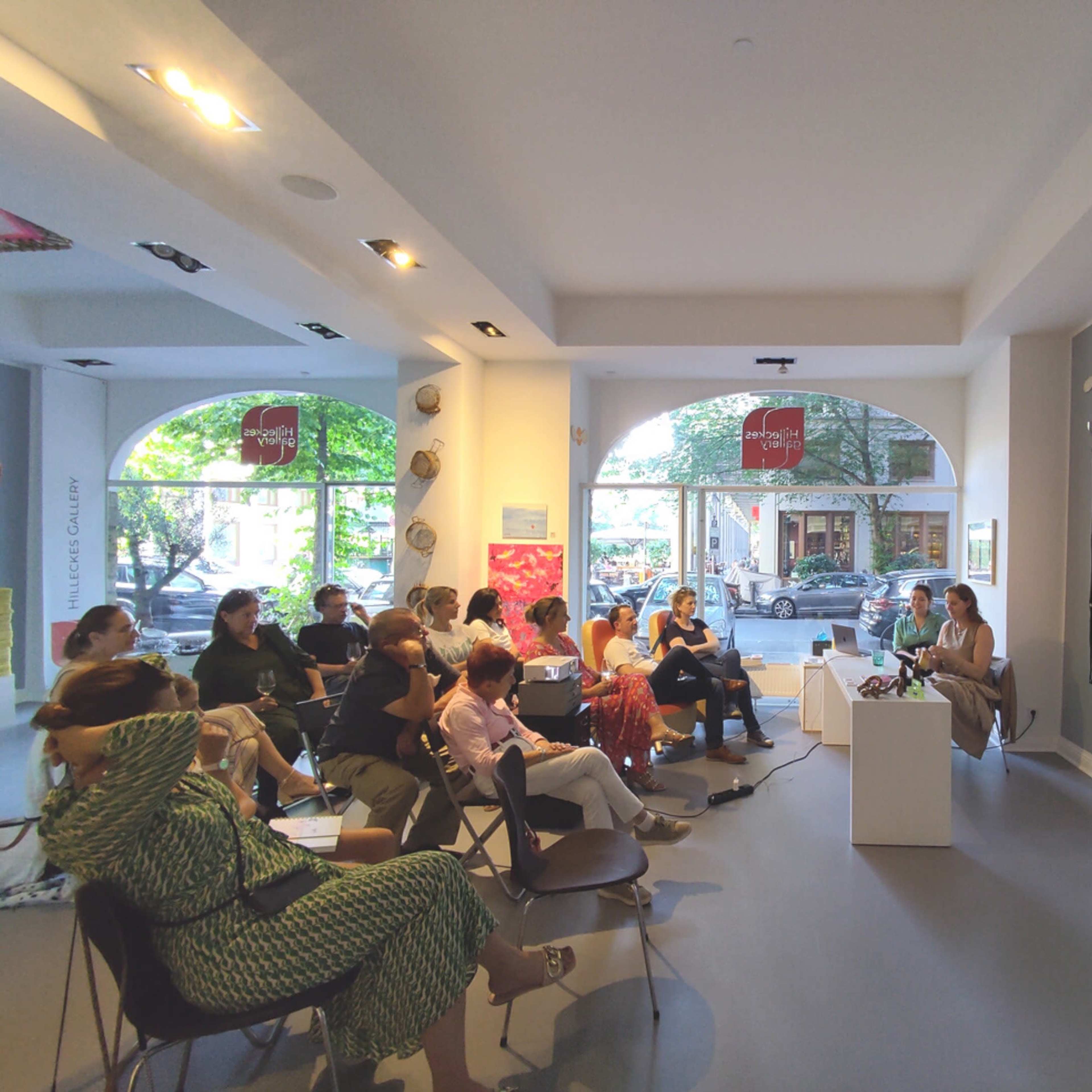 A group of people is seated in a gallery, watching a presentation that features a video projected on a wall.