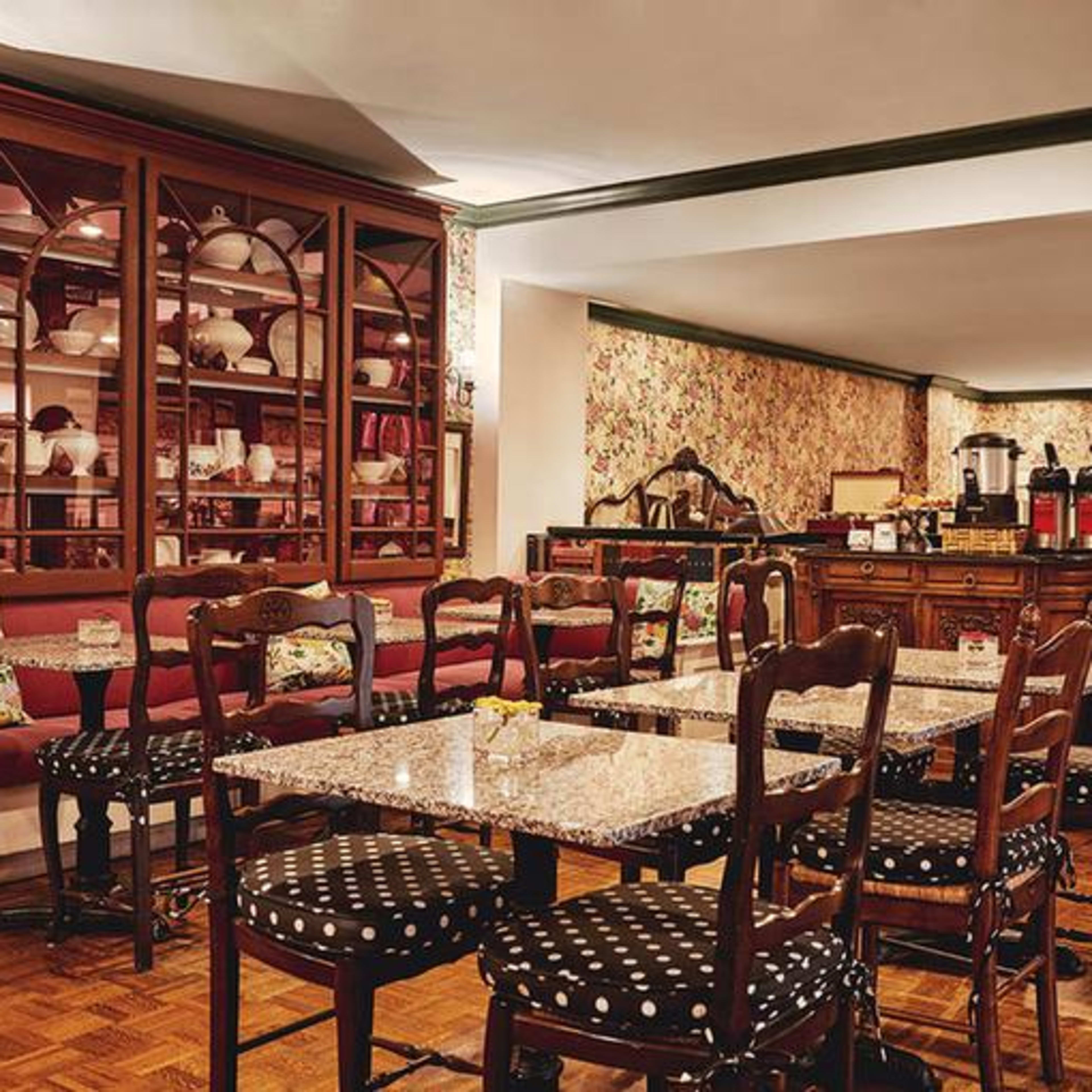 A cozy dining area with wooden tables and chairs, a floral wallpaper backdrop, and a display cabinet filled with dishware.