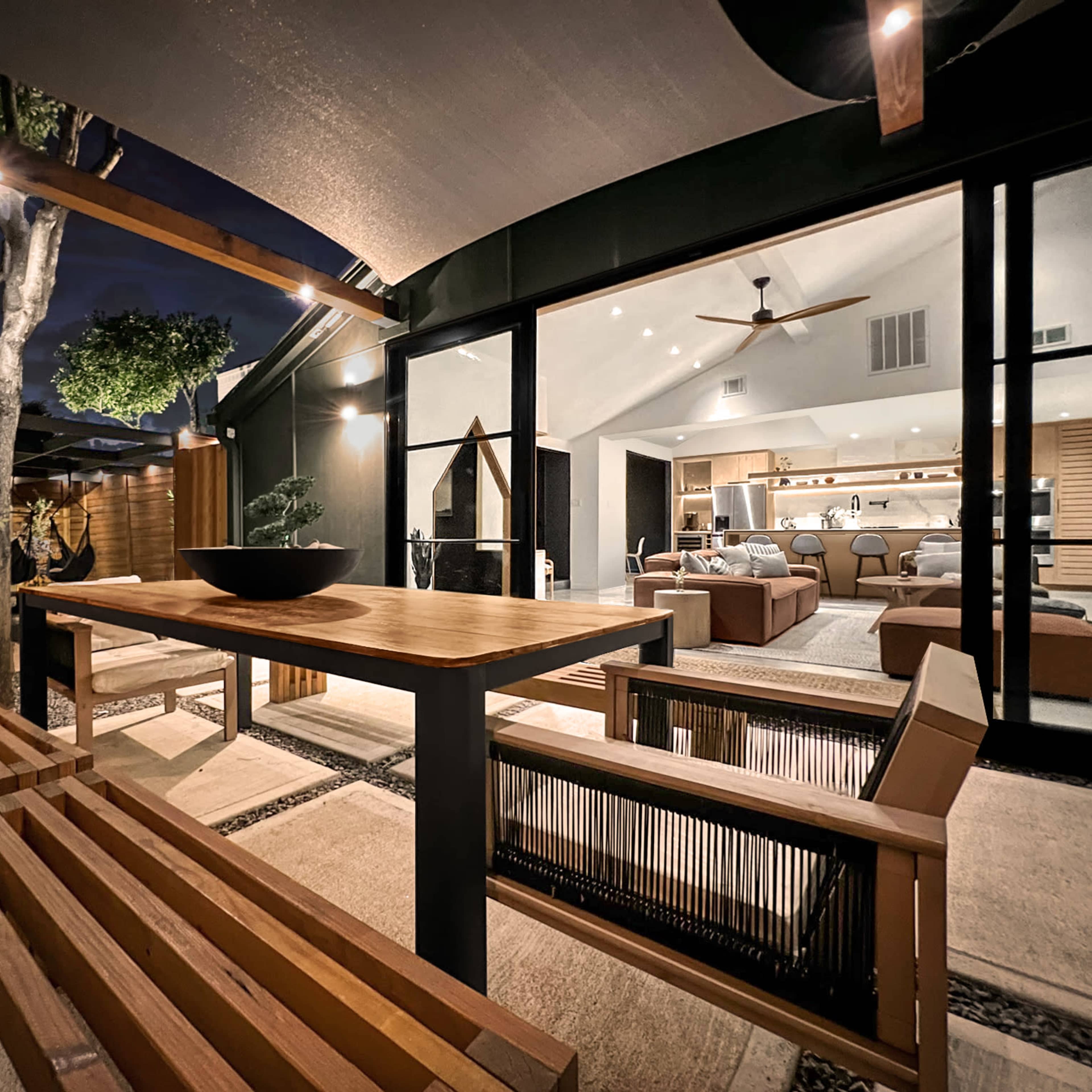 A wooden dining table and benches are positioned under a covered patio, adjacent to a modern interior space with large glass doors illuminated by indoor lights.