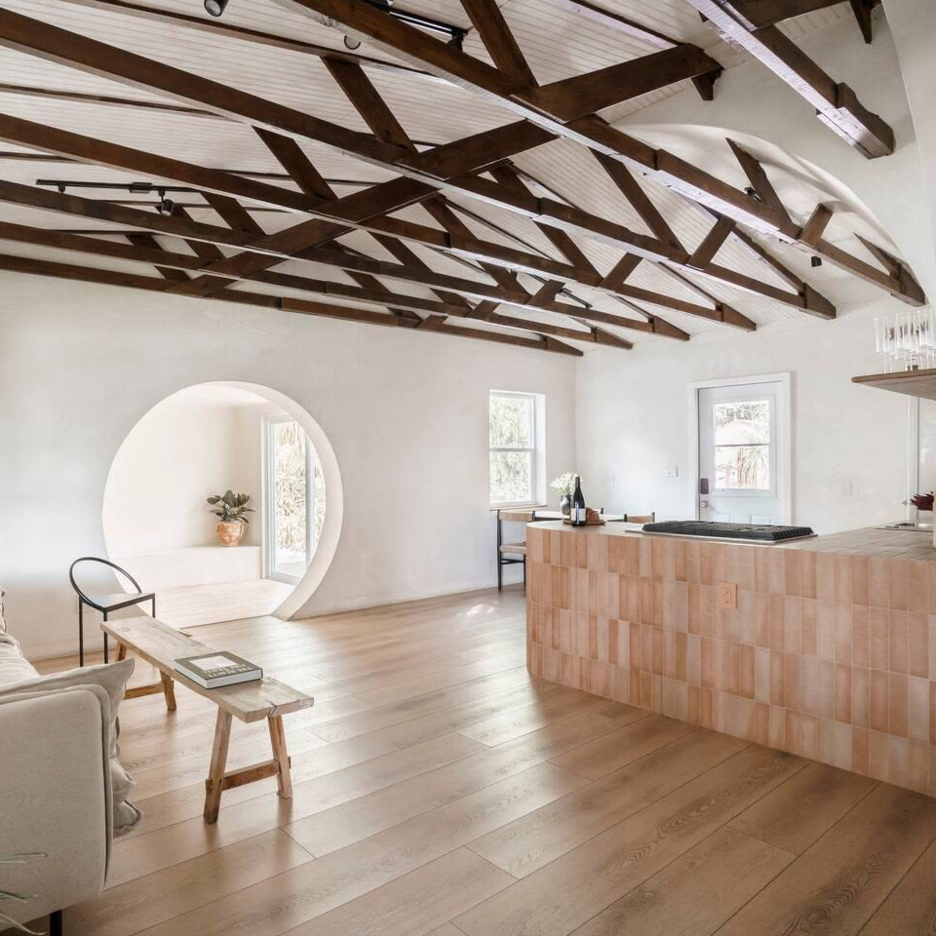 A modern, open living space featuring a light-colored sofa, a wooden dining table, and a round architectural opening leading to another room, all beneath exposed wooden beams.