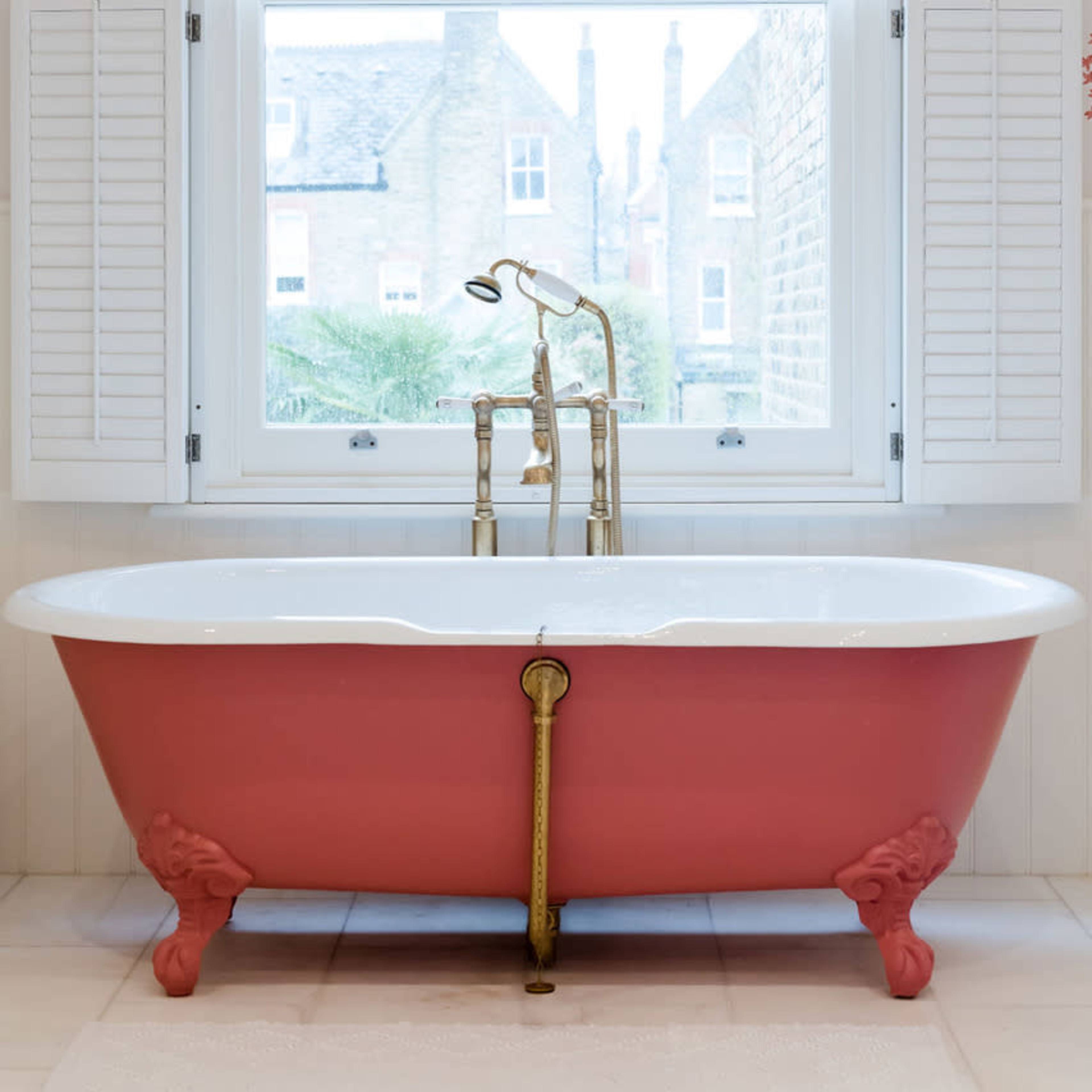 A pink and white clawfoot bathtub is positioned beneath a window with shuttered blinds, featuring a chandelier above and floral decor on the walls.