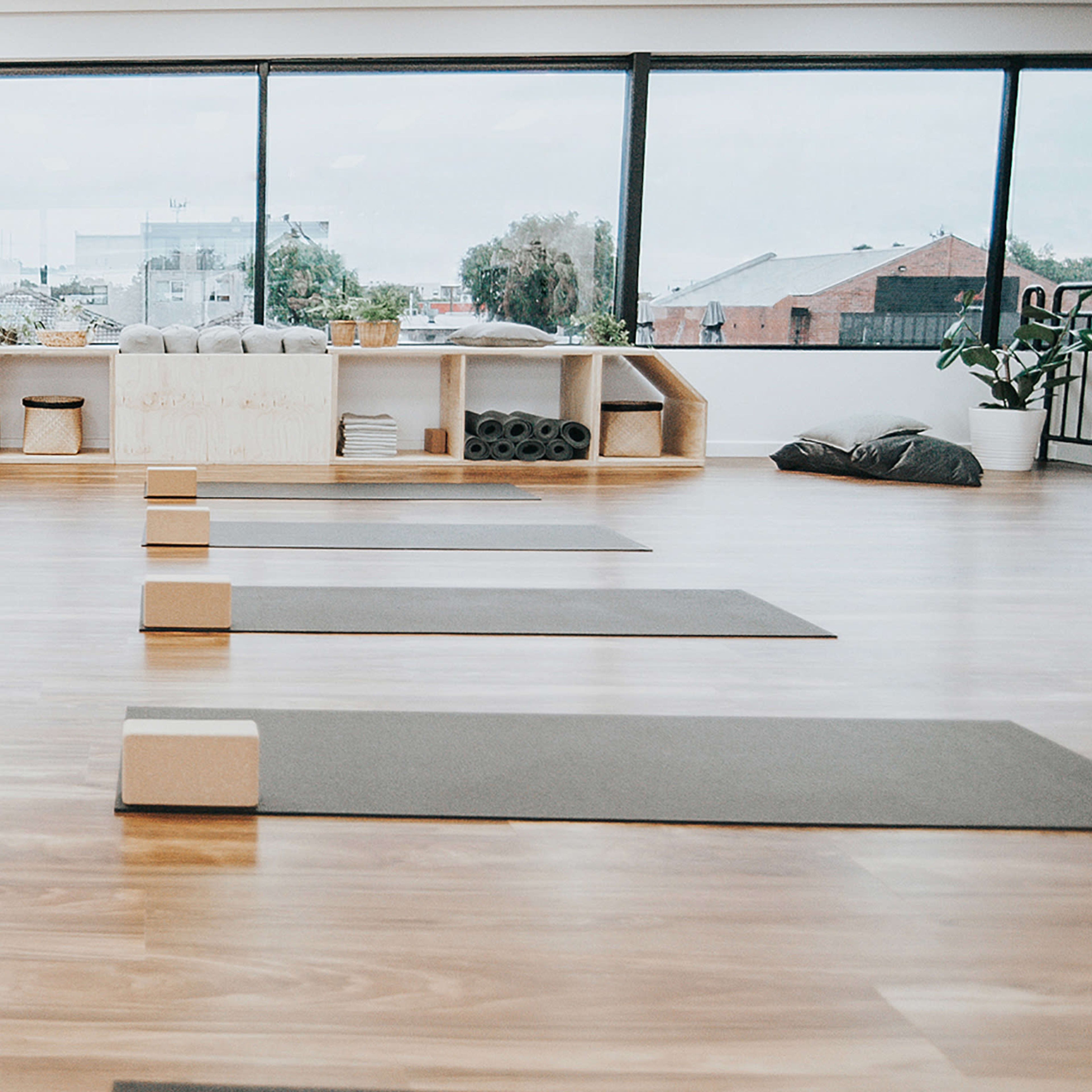 A spacious yoga studio with several yoga mats arranged on a wooden floor, blocks placed on the mats, and a seating area with storage shelves in the background.