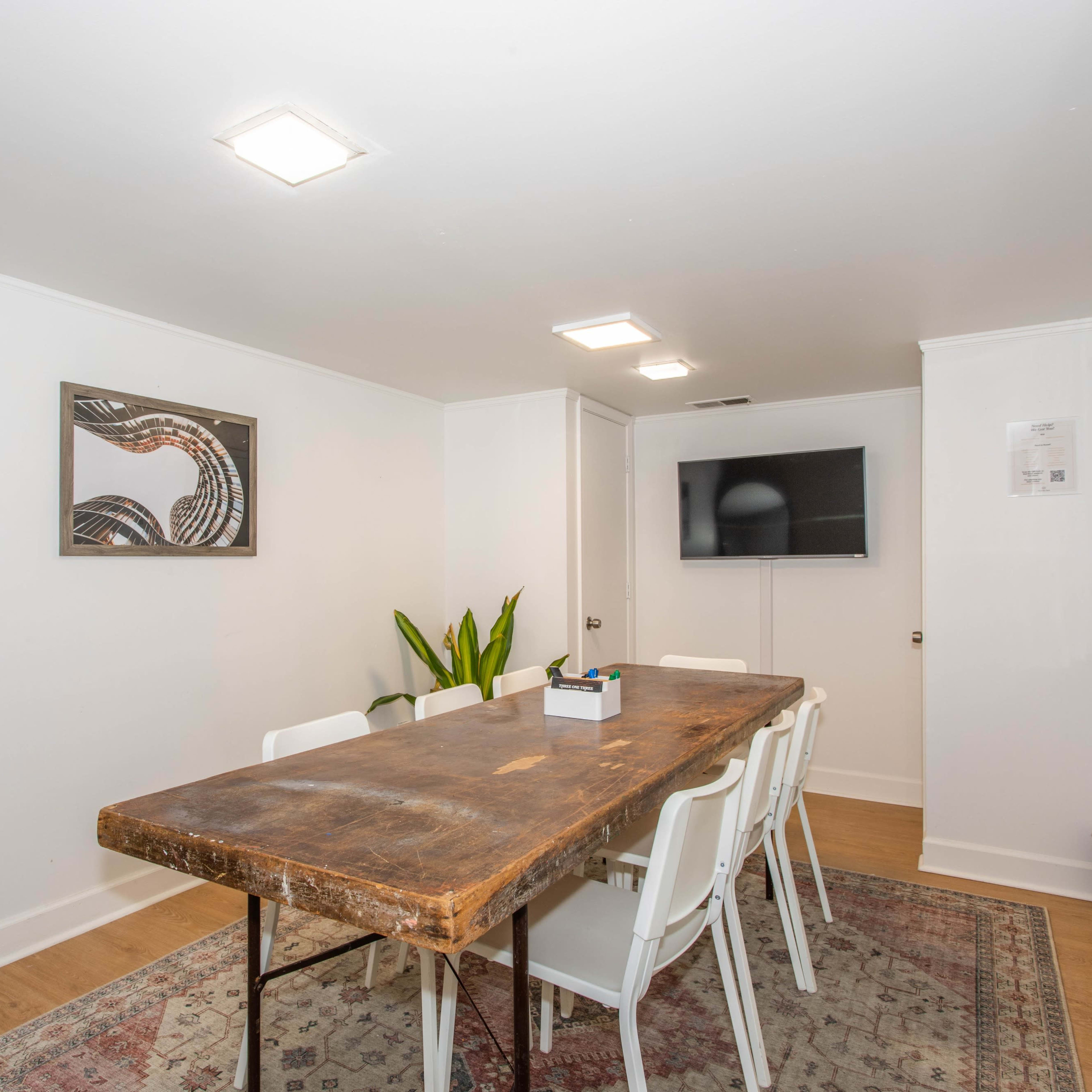 A spacious room features a long wooden table surrounded by white chairs, with a wall-mounted television and a large mirror.