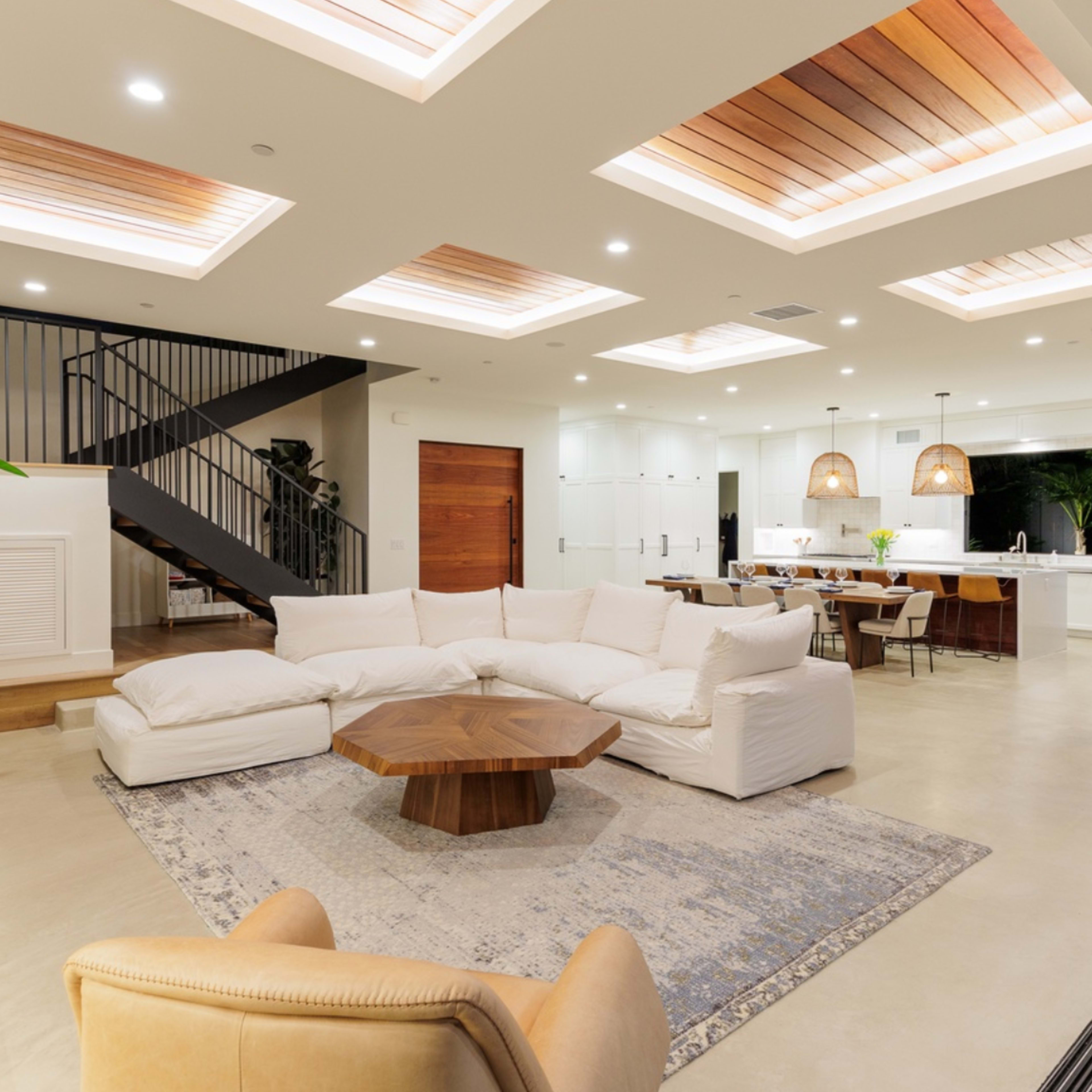 A modern living room with a white sectional sofa, a wooden coffee table, and a staircase leading to an upper level, adjacent to a dining area and kitchen.