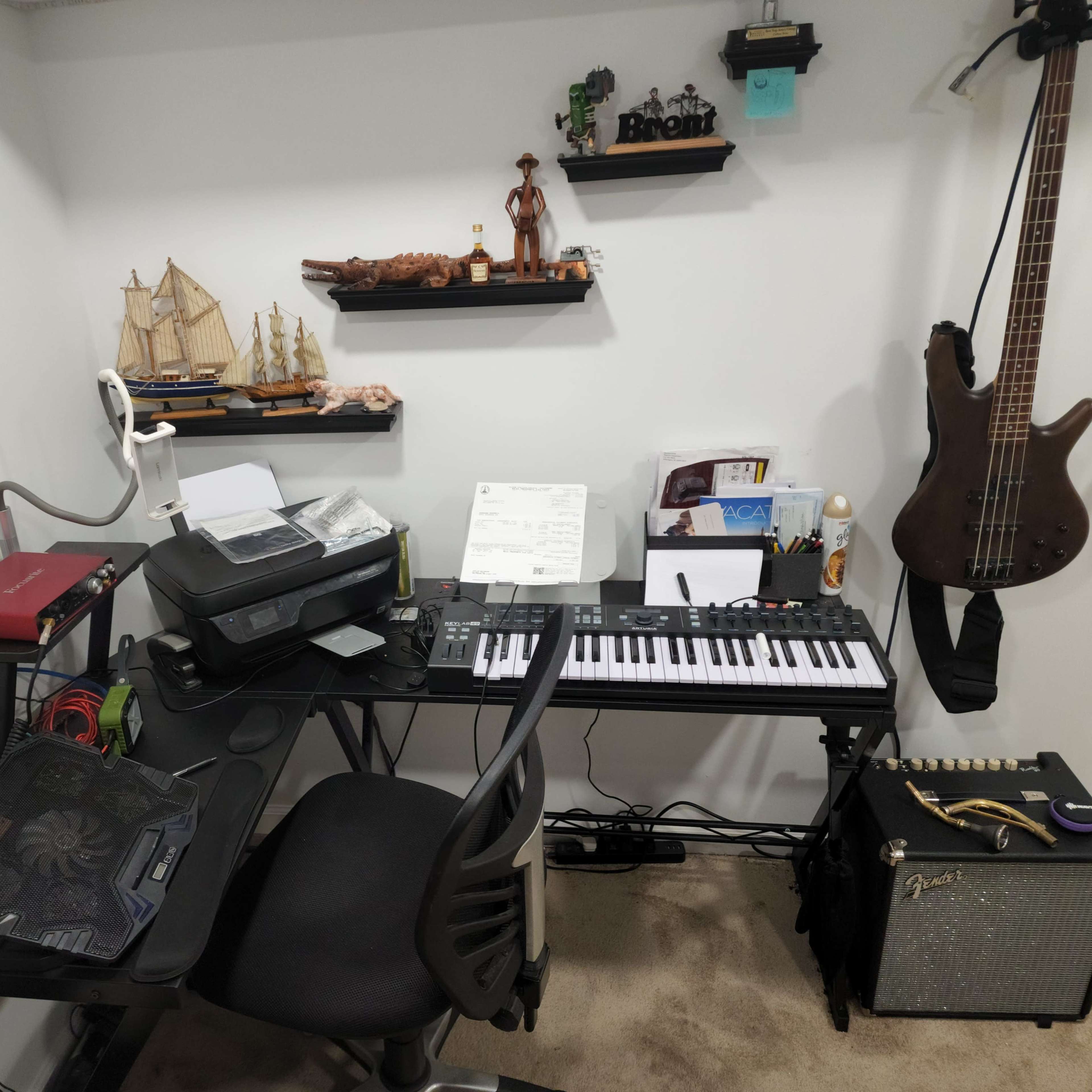 A corner workspace featuring a keyboard, sheets of music, a printer, and a guitar, with shelves displaying decorative items and a diploma on the wall.