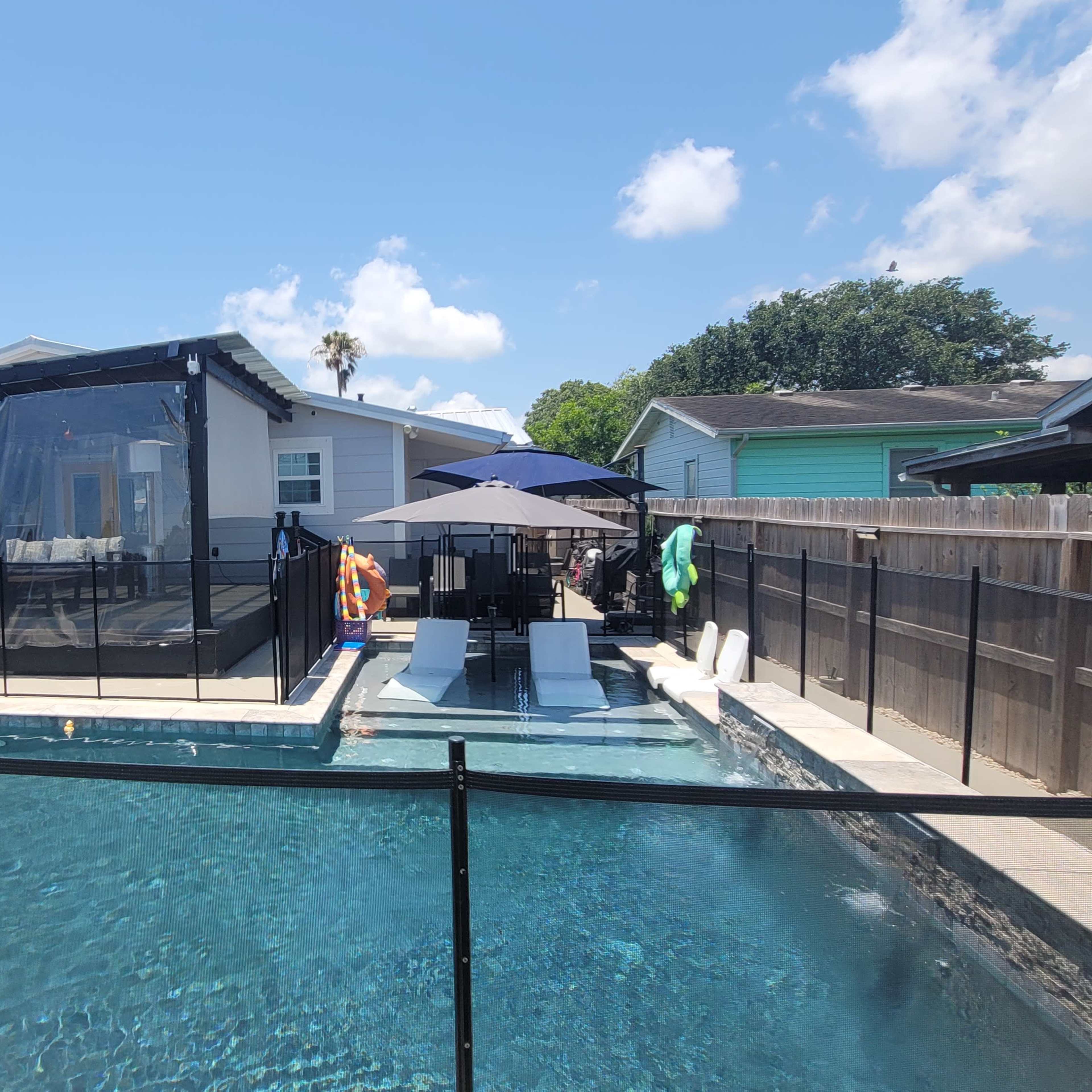 A backyard swimming pool enclosed by a black safety fence, with loungers and an umbrella on a deck next to a house.