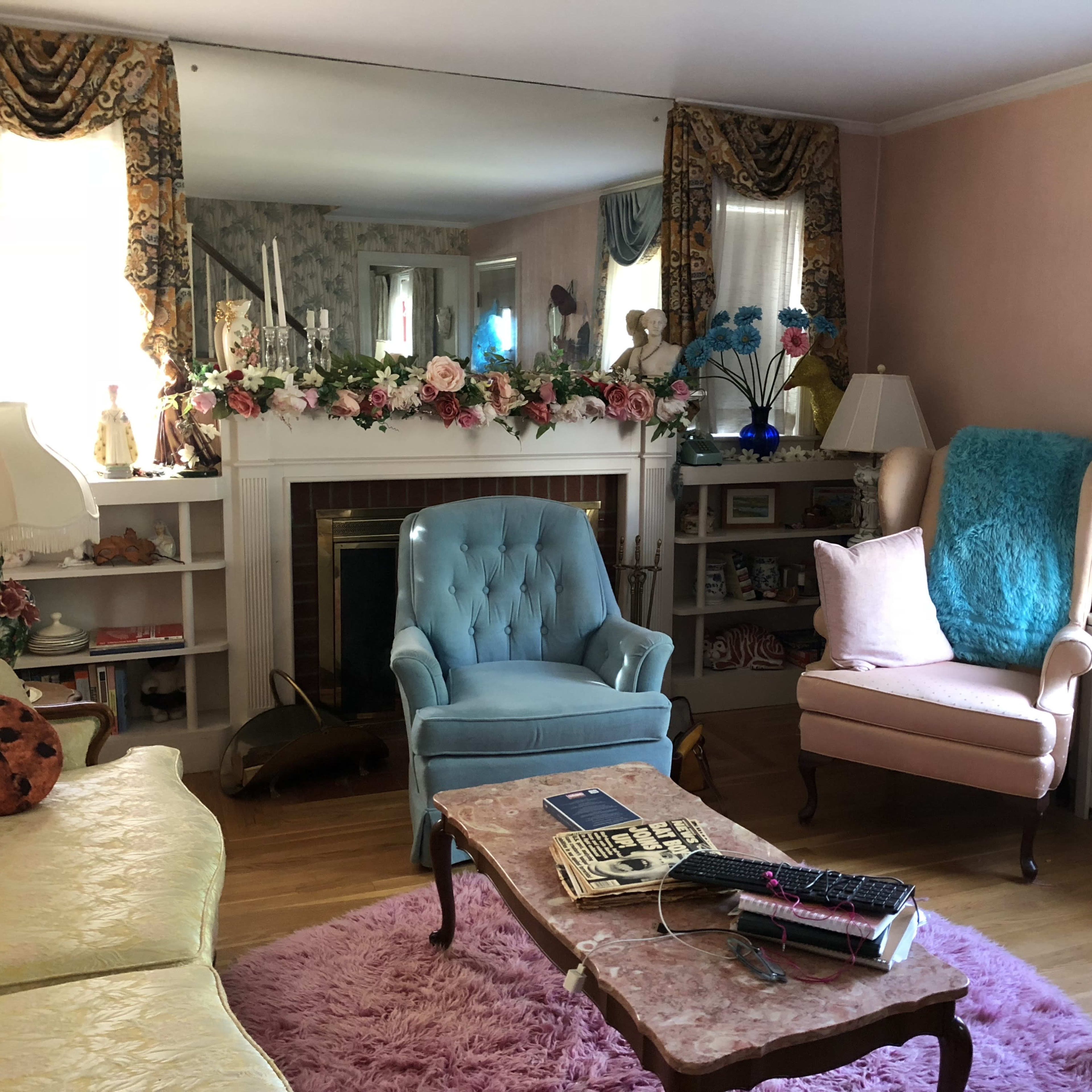 The room features a pastel color scheme with a pink rug, two upholstered chairs, a floral arrangement on the mantel, and a large mirror reflecting the space.