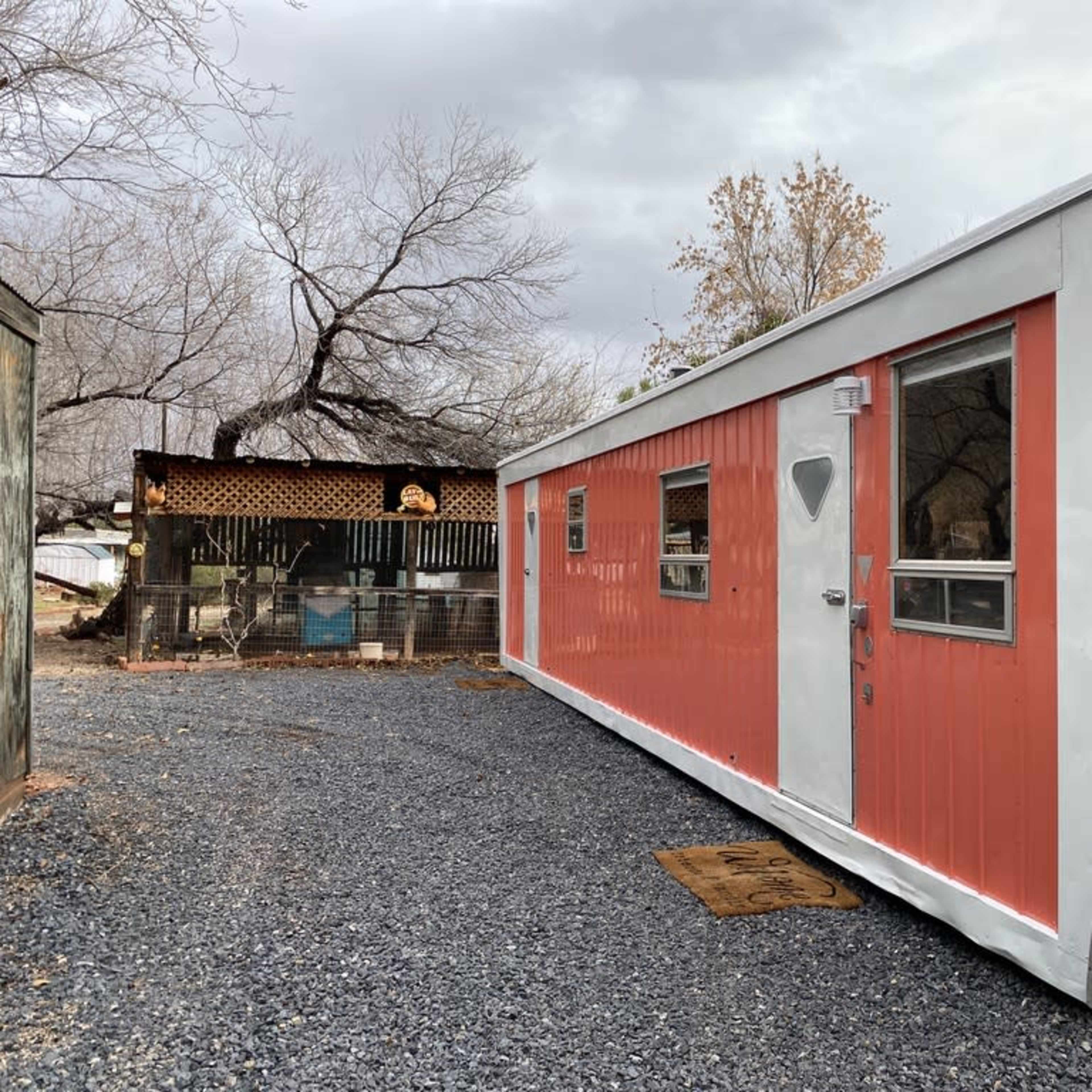 A retro-style mobile home with orange and white siding is parked on a gravel driveway next to a rustic structure in a treed outdoor setting.
