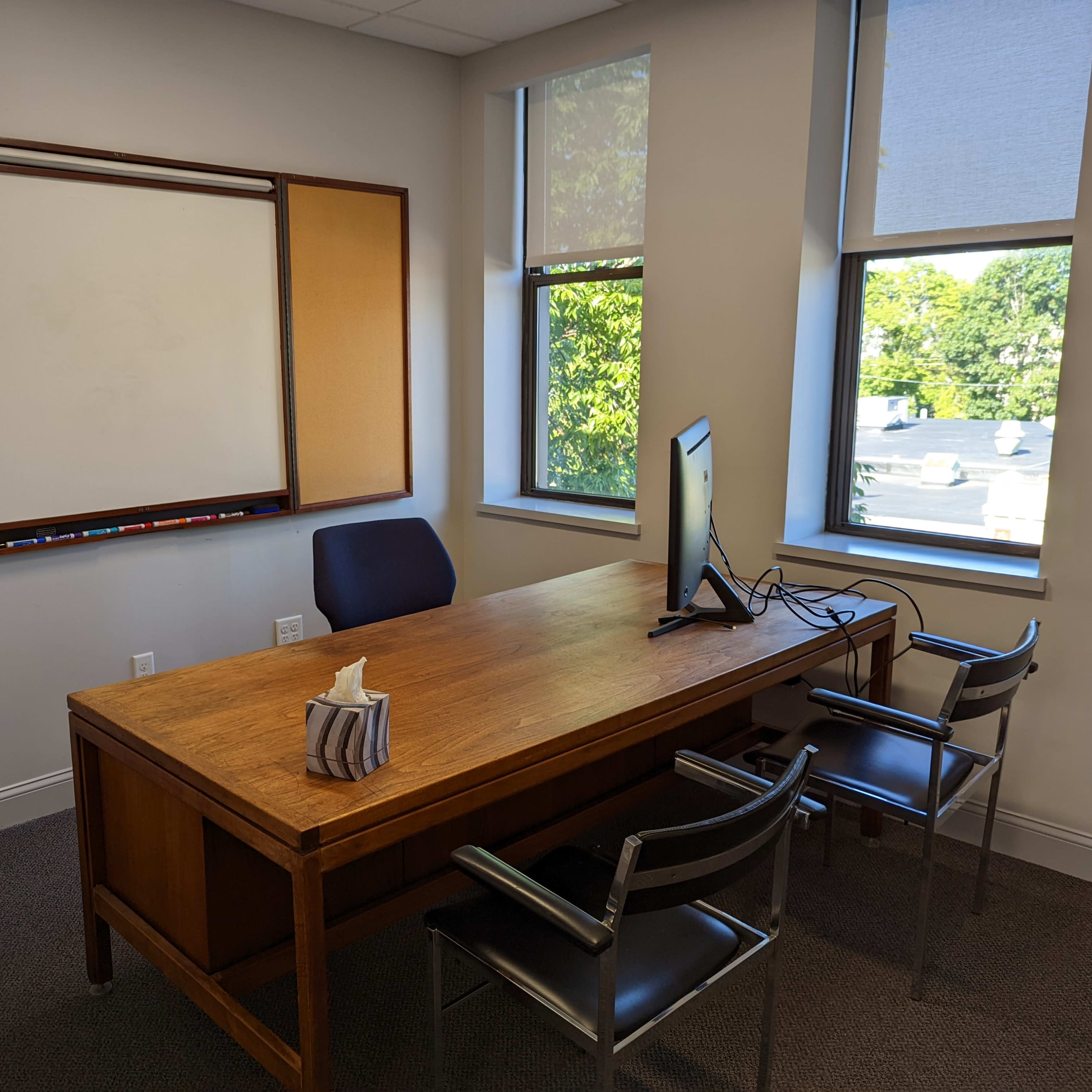 A small, well-lit meeting room features a wooden table, a chair, a computer monitor, and a whiteboard.