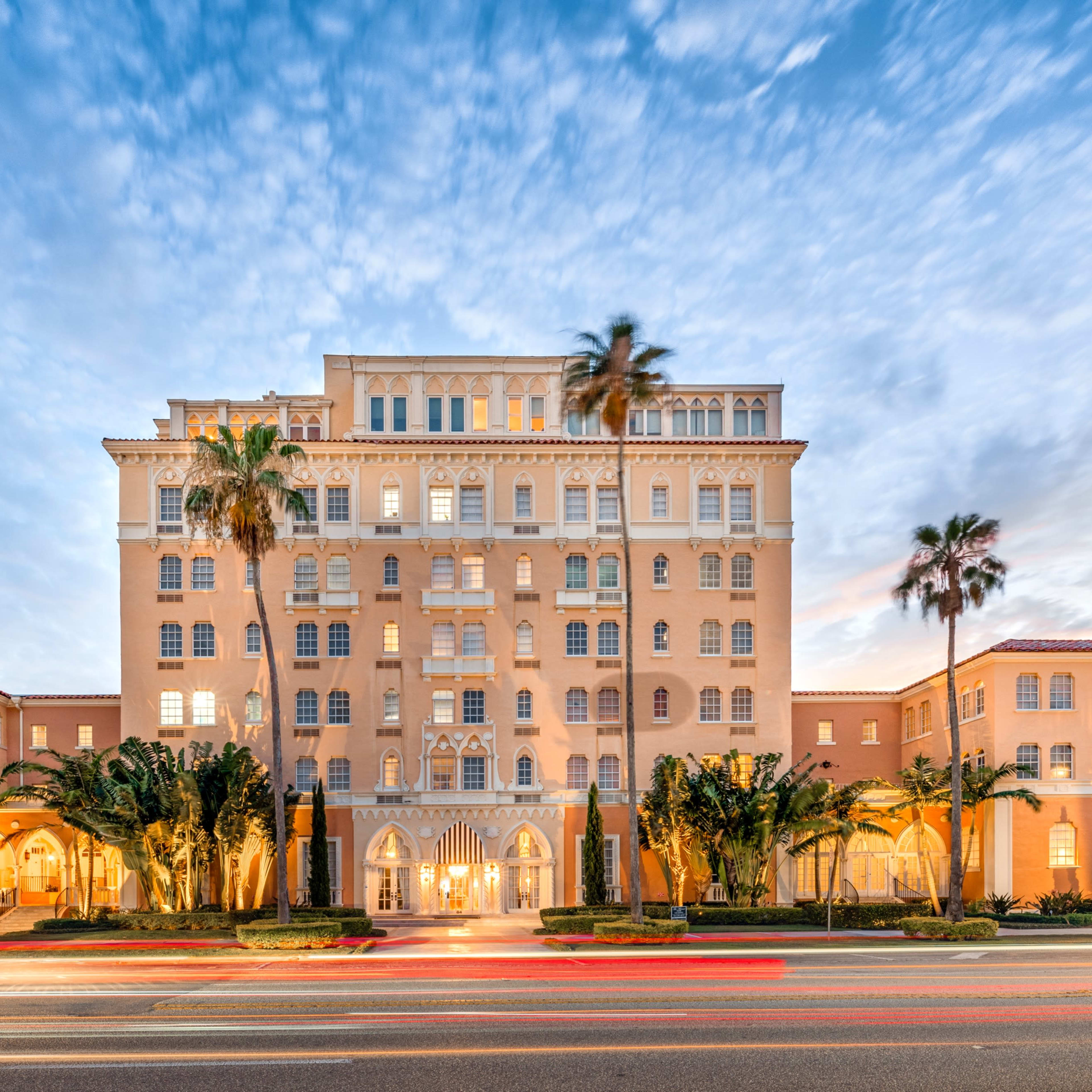 A large, pale pink hotel with multiple stories and palm trees stands against a blue sky with scattered clouds.