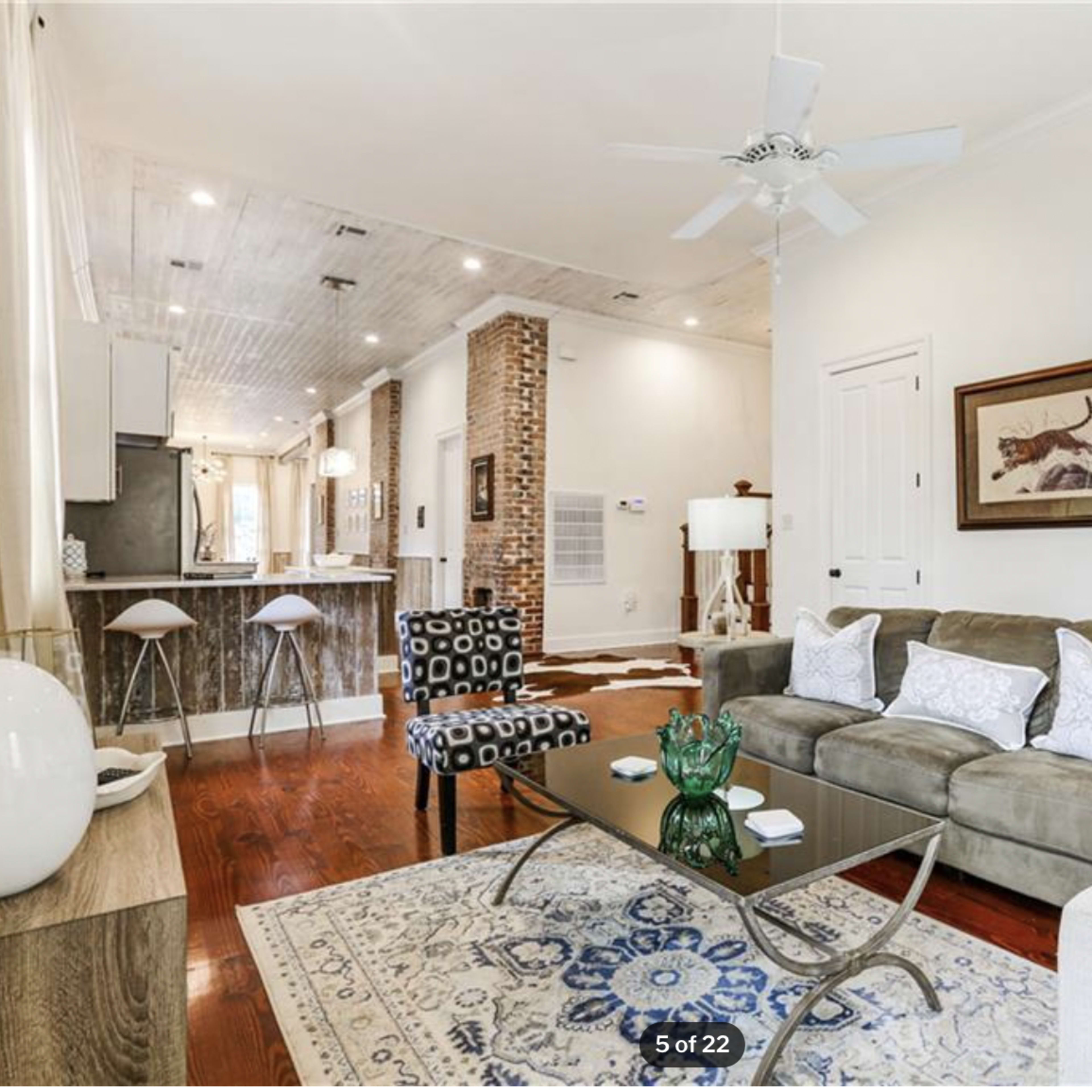 A living room with a beige sofa, patterned chairs, a coffee table, and a kitchen area in the background featuring bar stools and exposed brick.