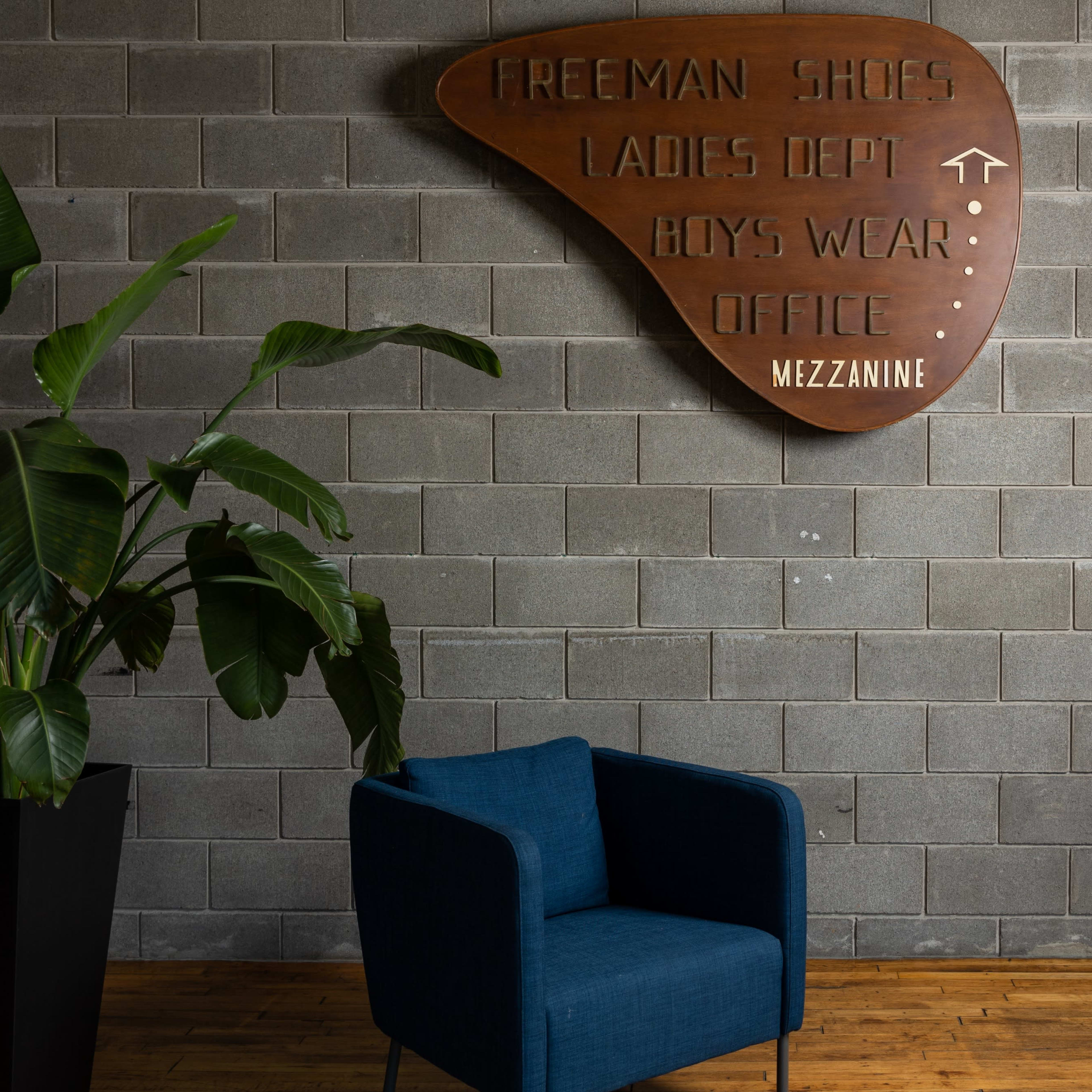 A blue armchair is positioned in front of a textured gray wall featuring a large, wooden sign with directional information about various store departments.