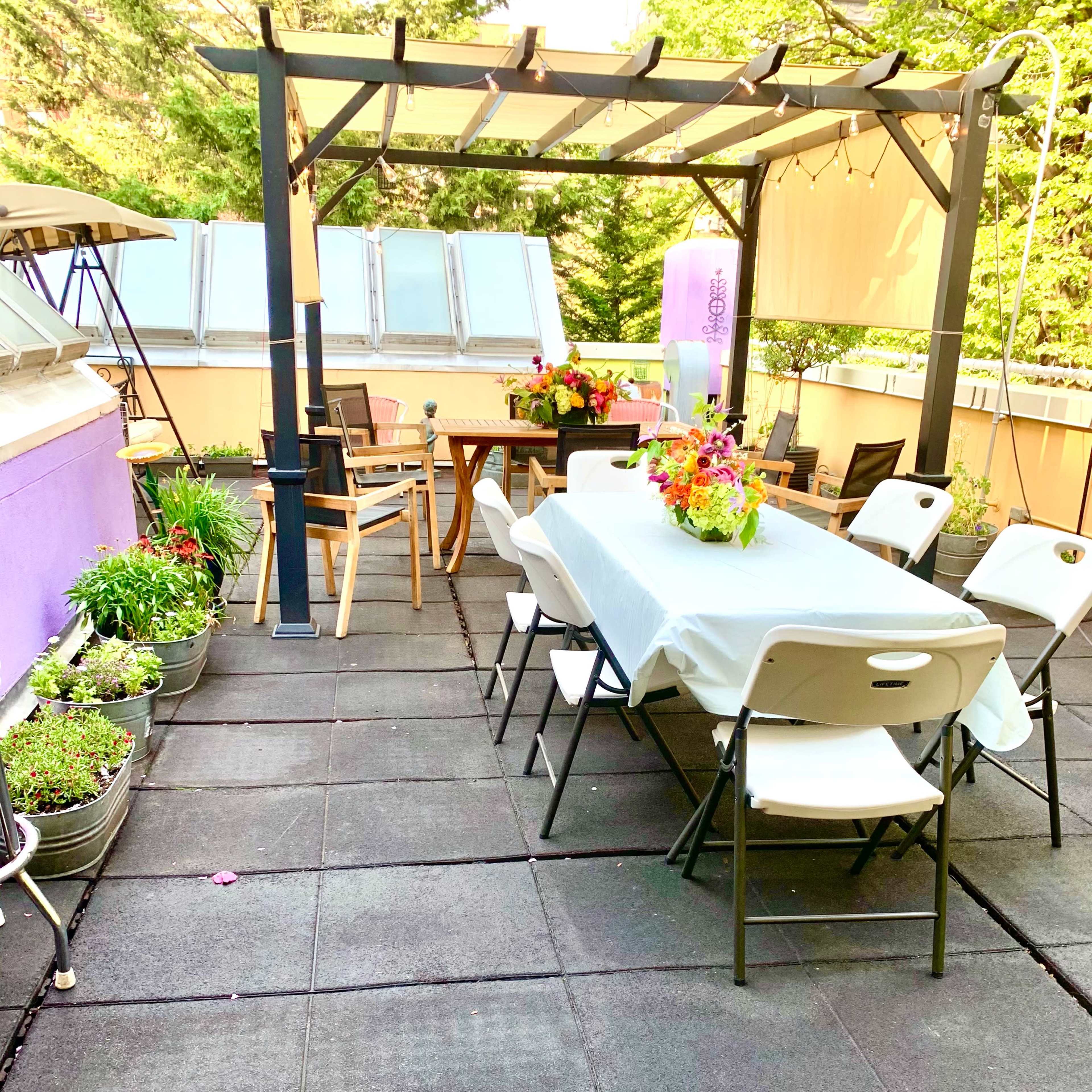 A patio features a long table covered with a white tablecloth, surrounded by folding chairs, and adorned with flower arrangements, set under a pergola.