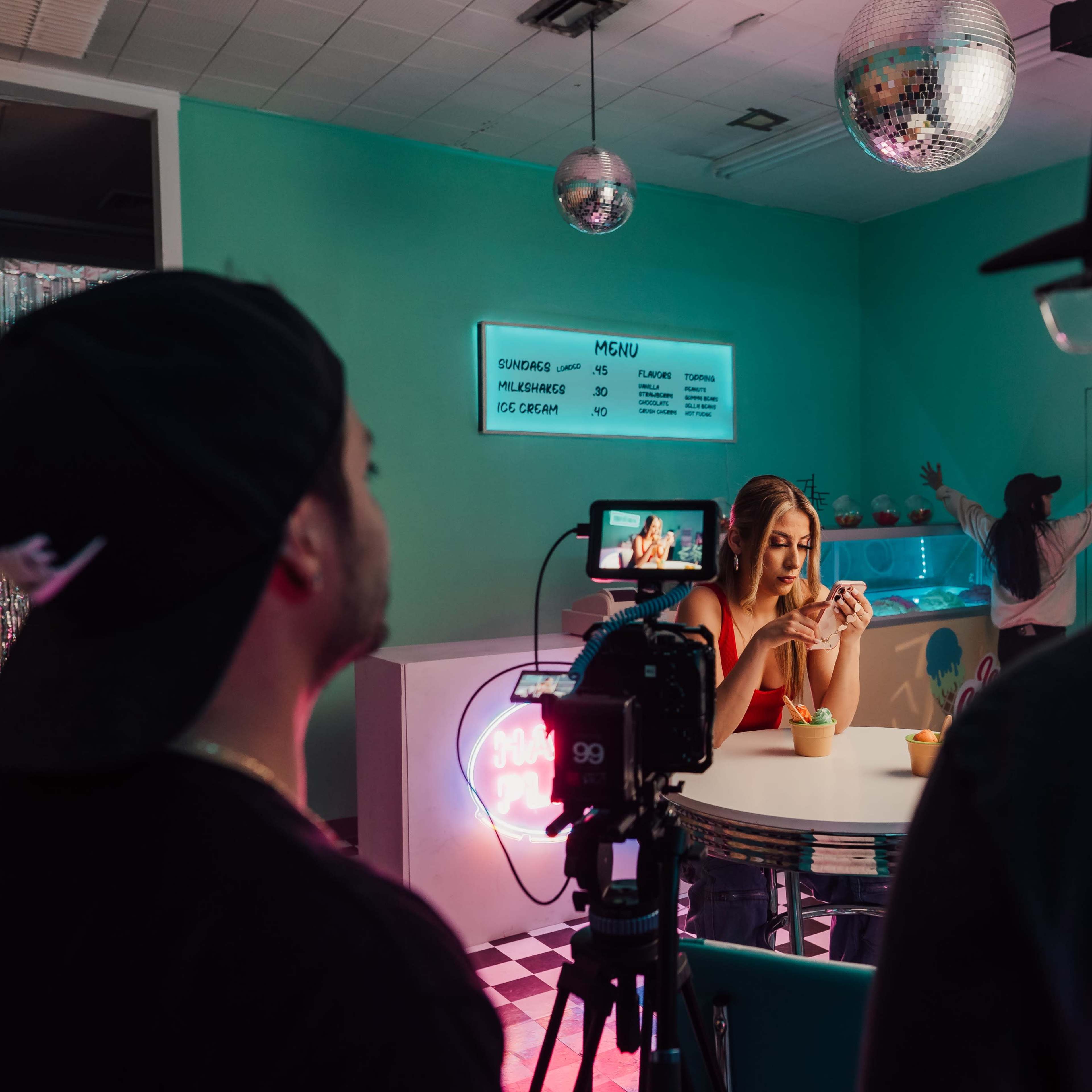 A woman sits at a table in a brightly colored diner while two people film her with a camera, and a person is visible in the background.