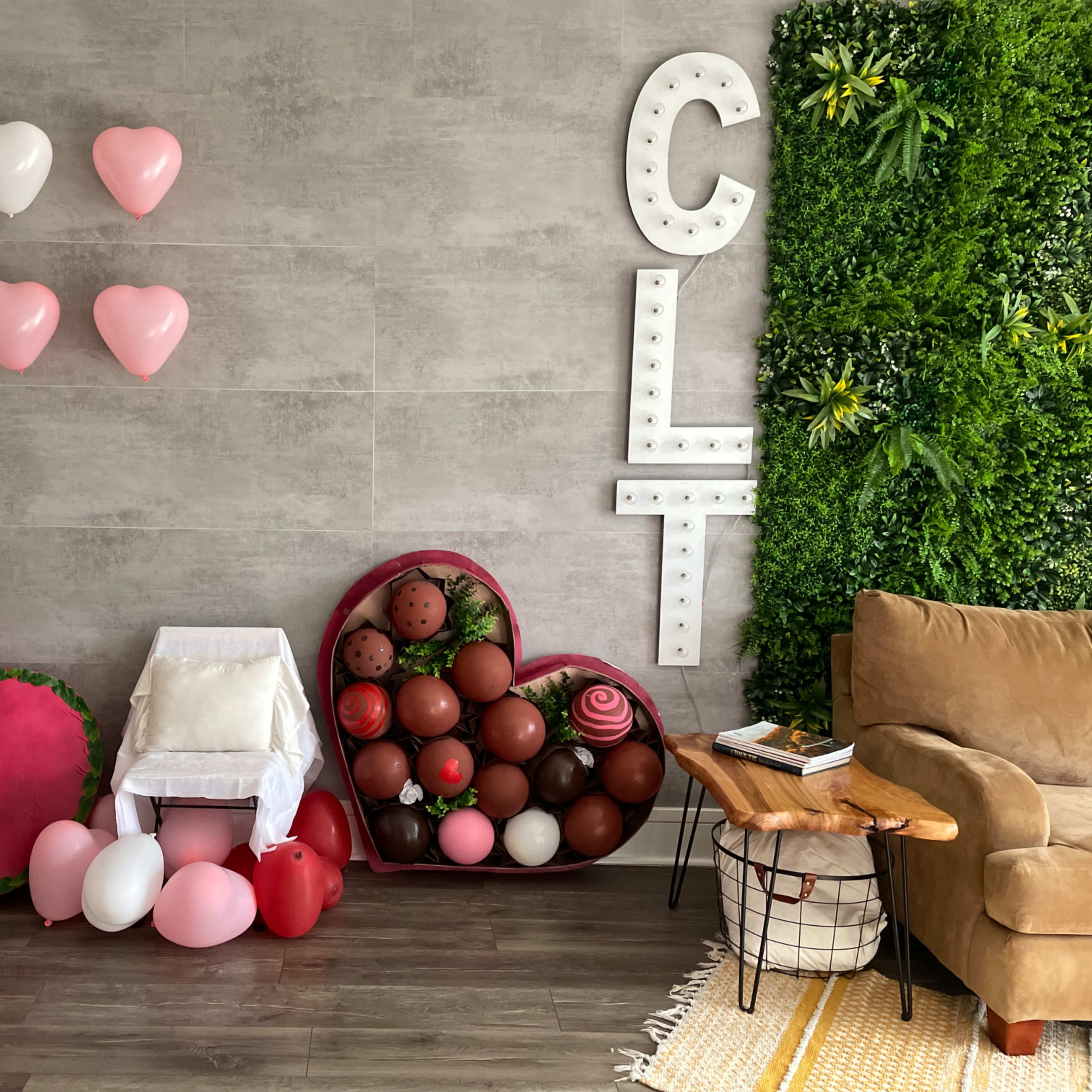 A decorated room with a gray wall featuring heart-shaped balloons, a large heart-shaped arrangement made of faux flowers and chocolates, and a cozy seating area with a brown sofa and a wooden side table.