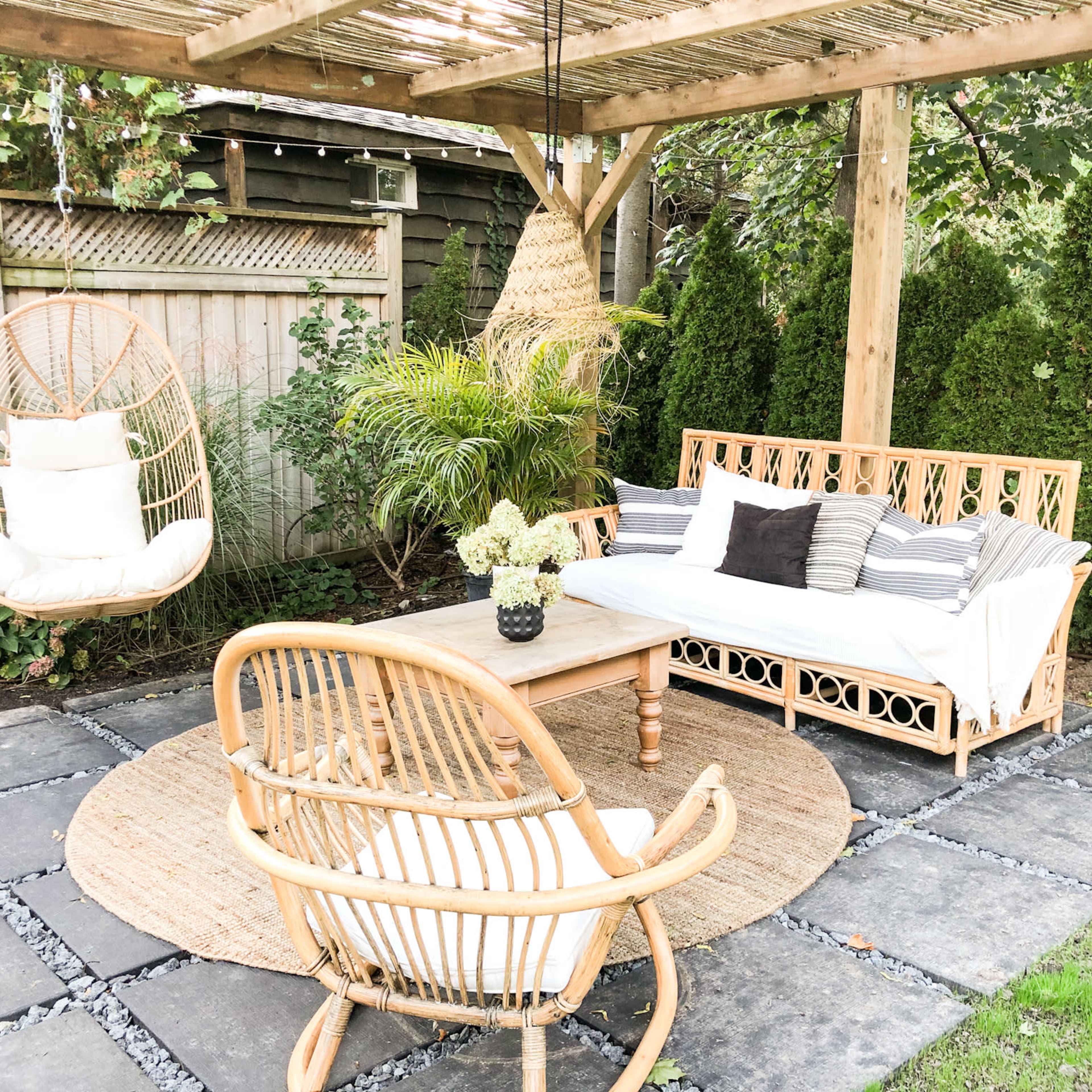 A shaded outdoor seating area with a rattan sofa, a circular rug, and a hanging chair surrounded by greenery.