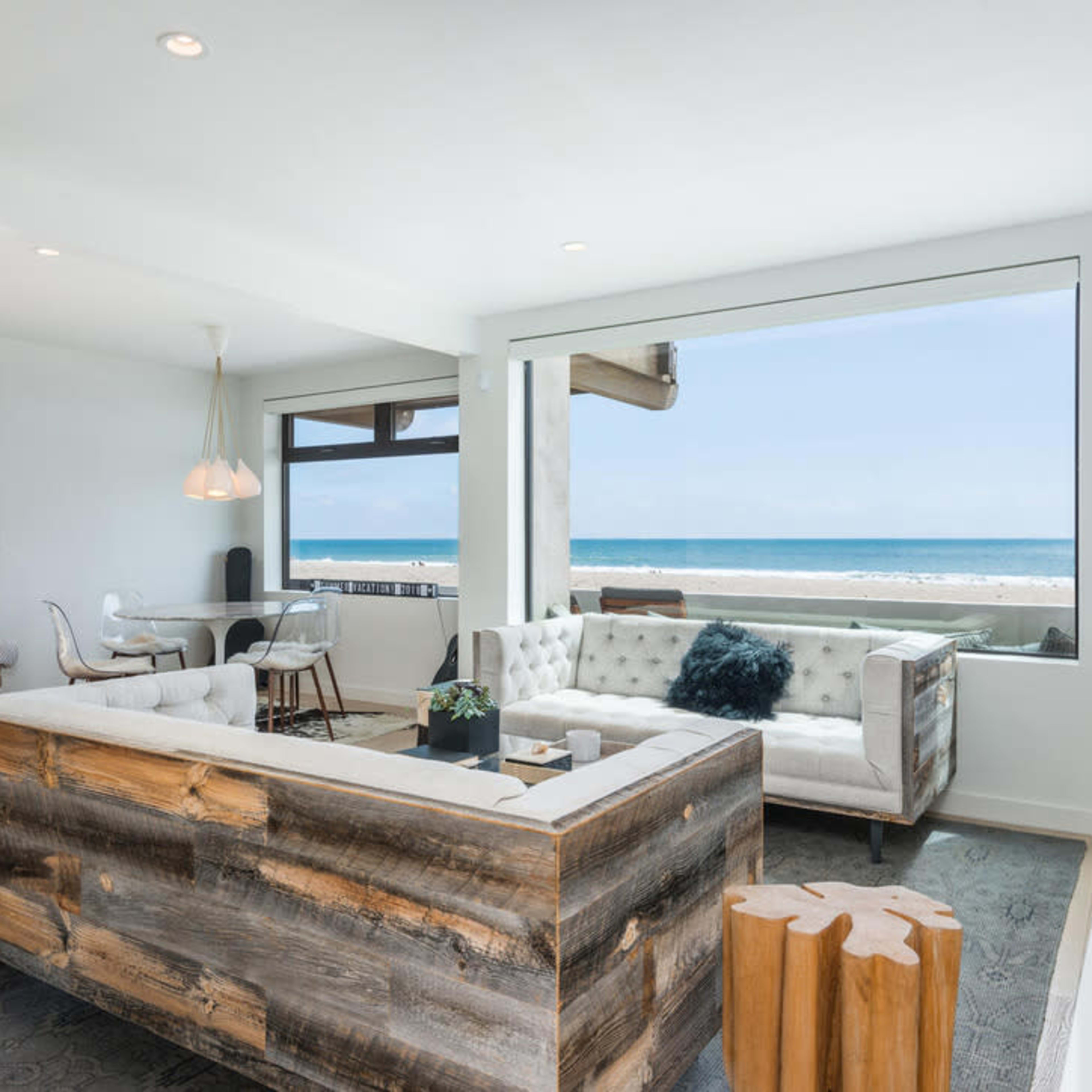 A modern living room features a wooden sofa, a small dining area, and large windows overlooking the ocean.