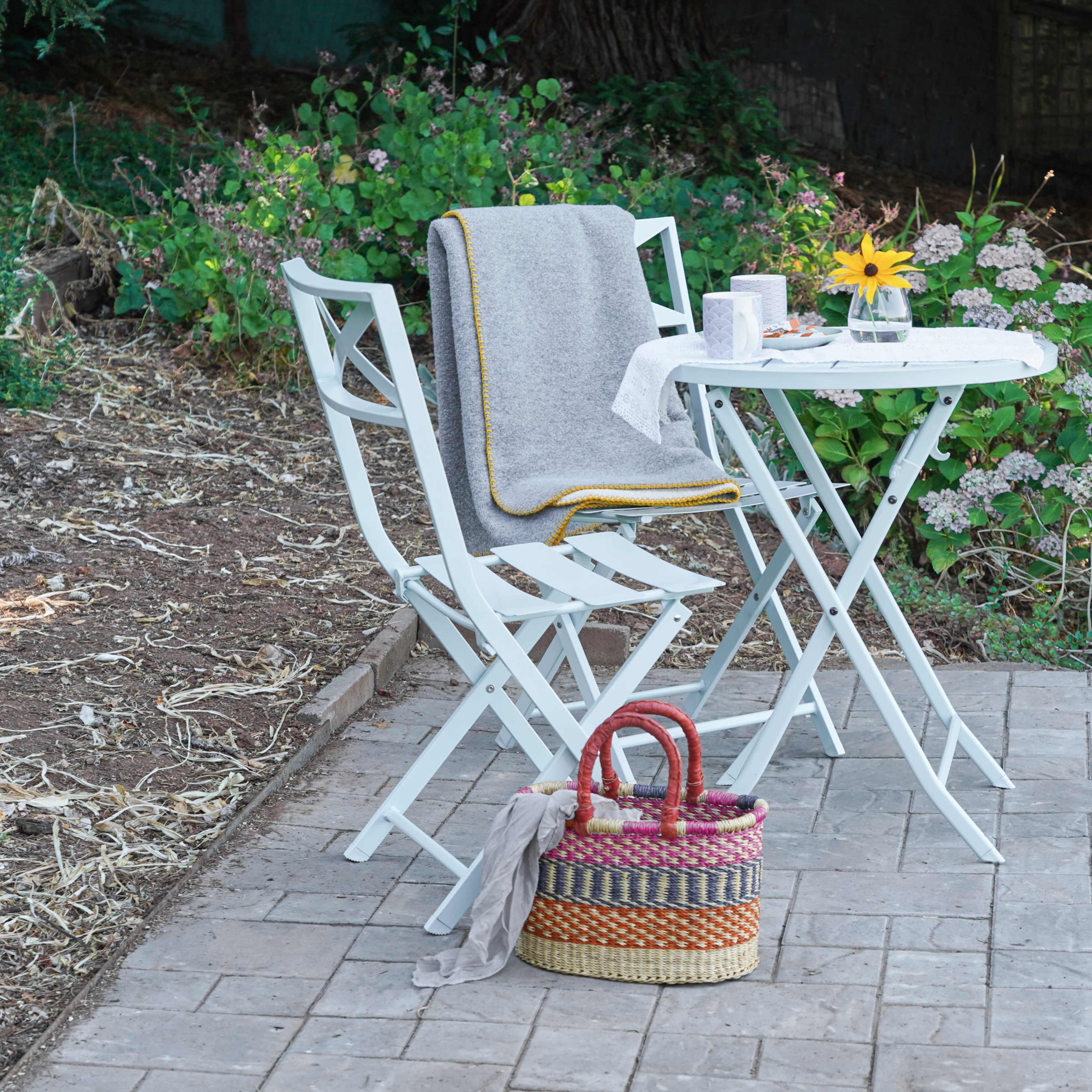A small outdoor table and two chairs are set beside a flowerbed, accompanied by a basket and a blanket draped over one of the chairs.