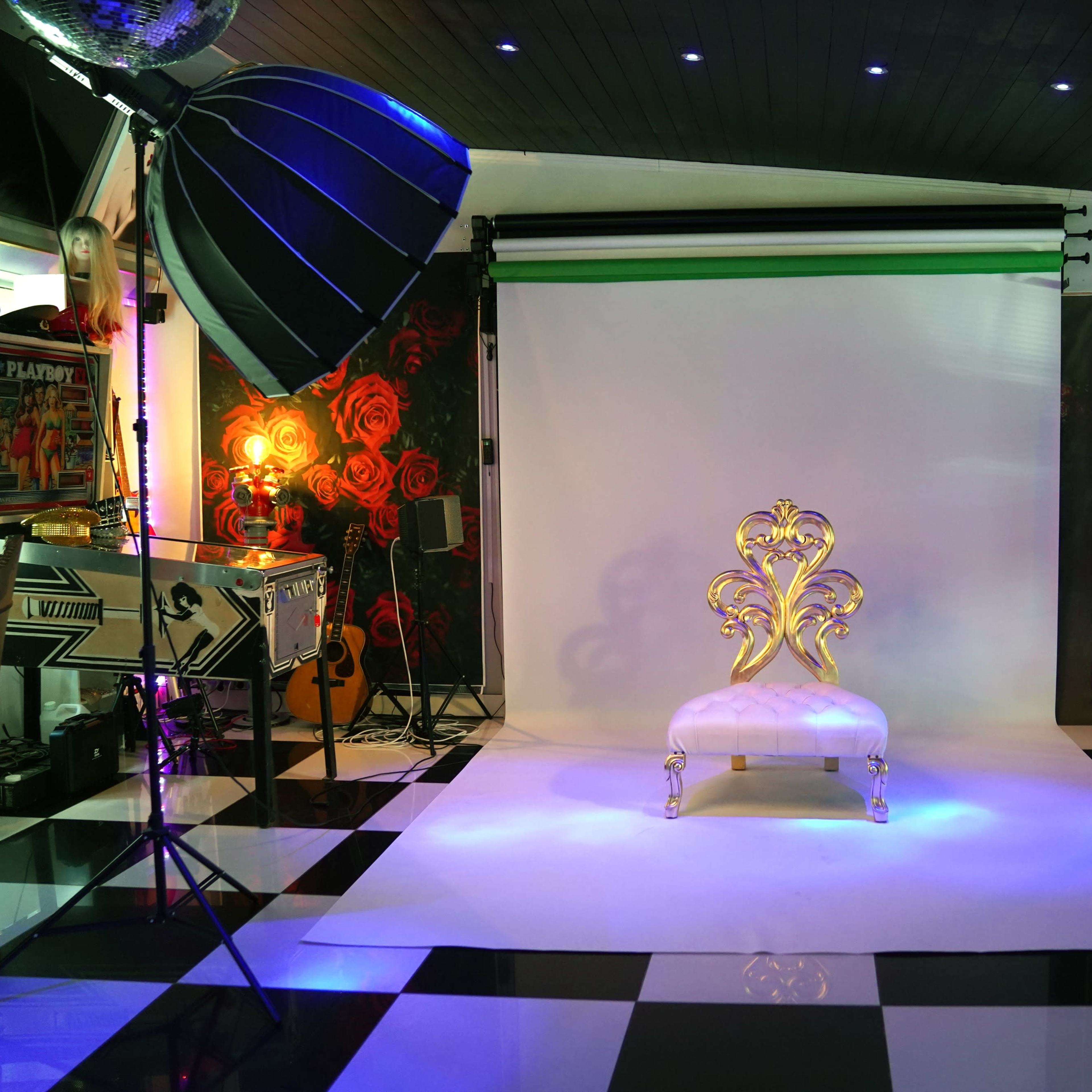 A photography studio with a white backdrop, a decorative gold chair, and various lighting equipment arranged around a black and white checkered floor.